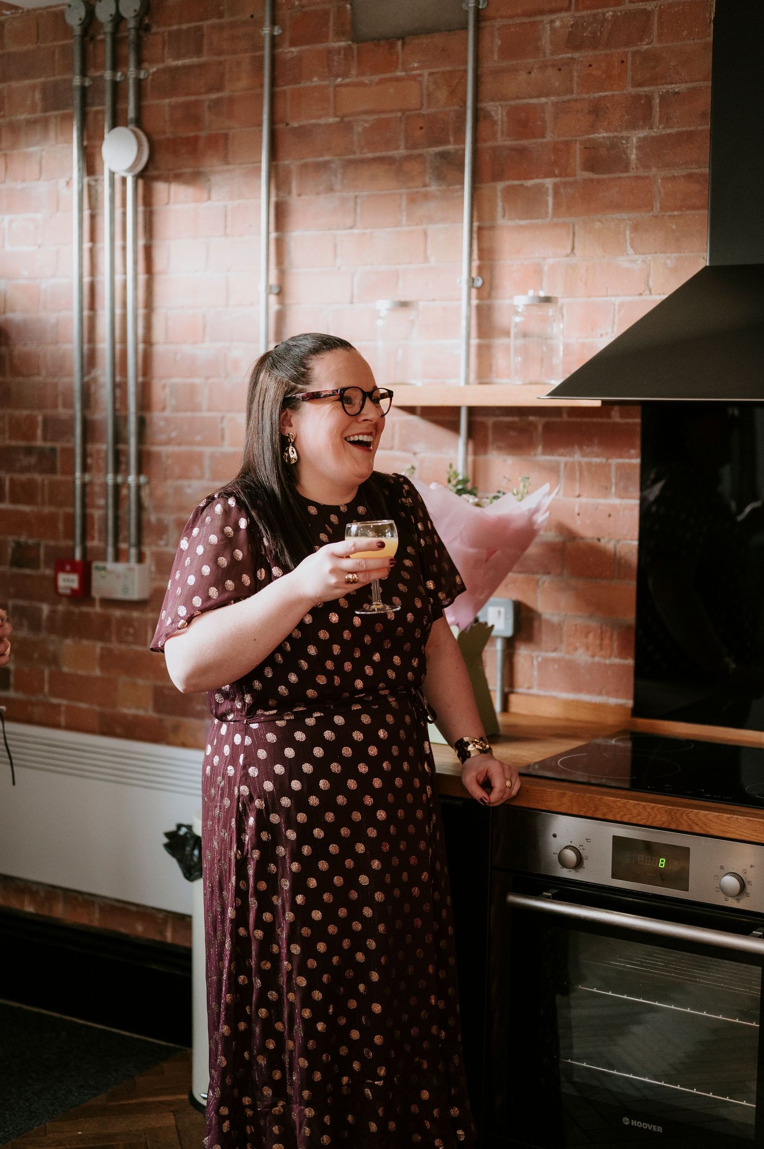 Woman in polka dot dress laughing while holding drink in industrial style kitchen.