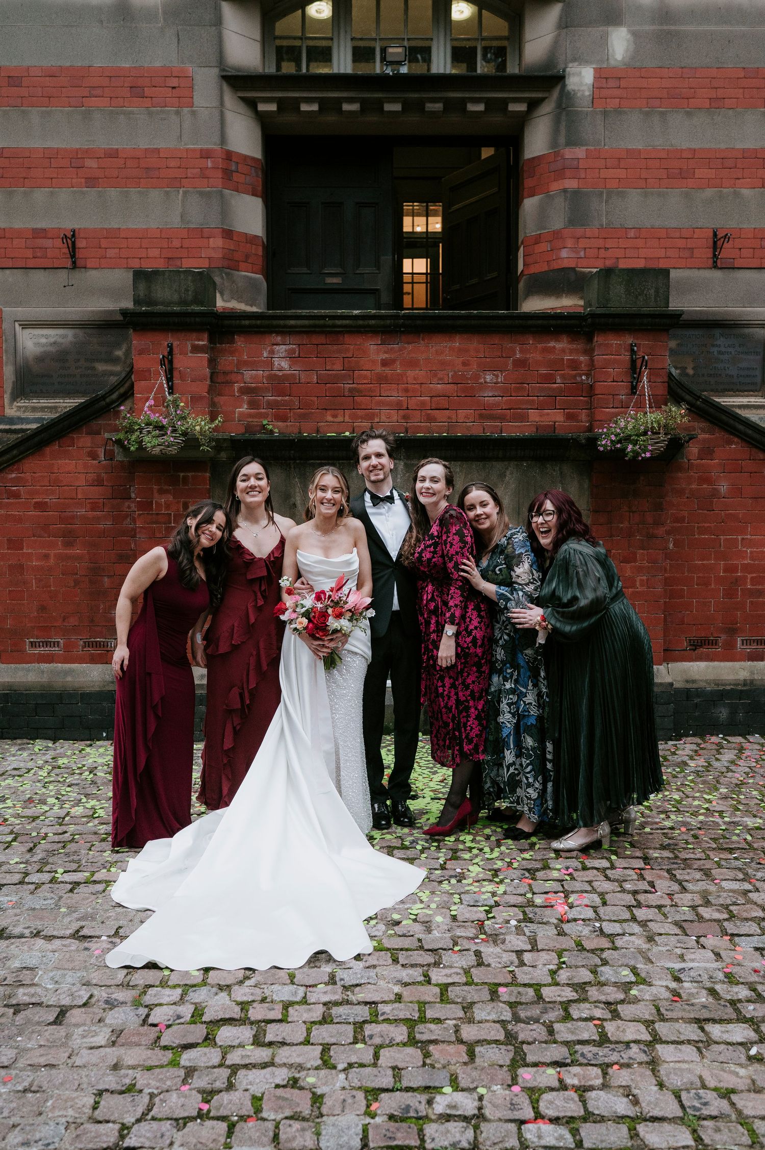 Wedding party in burgundy and black attire poses on cobblestone street against red brick building.