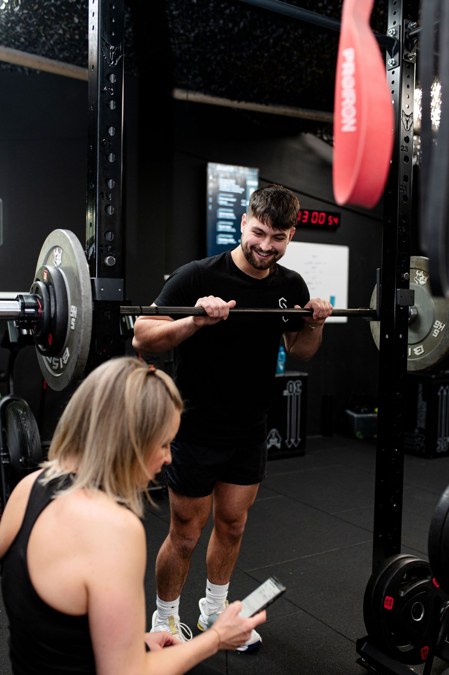 A trainer assists someone with barbell exercises in a dark gym setting with red weight plates visible.