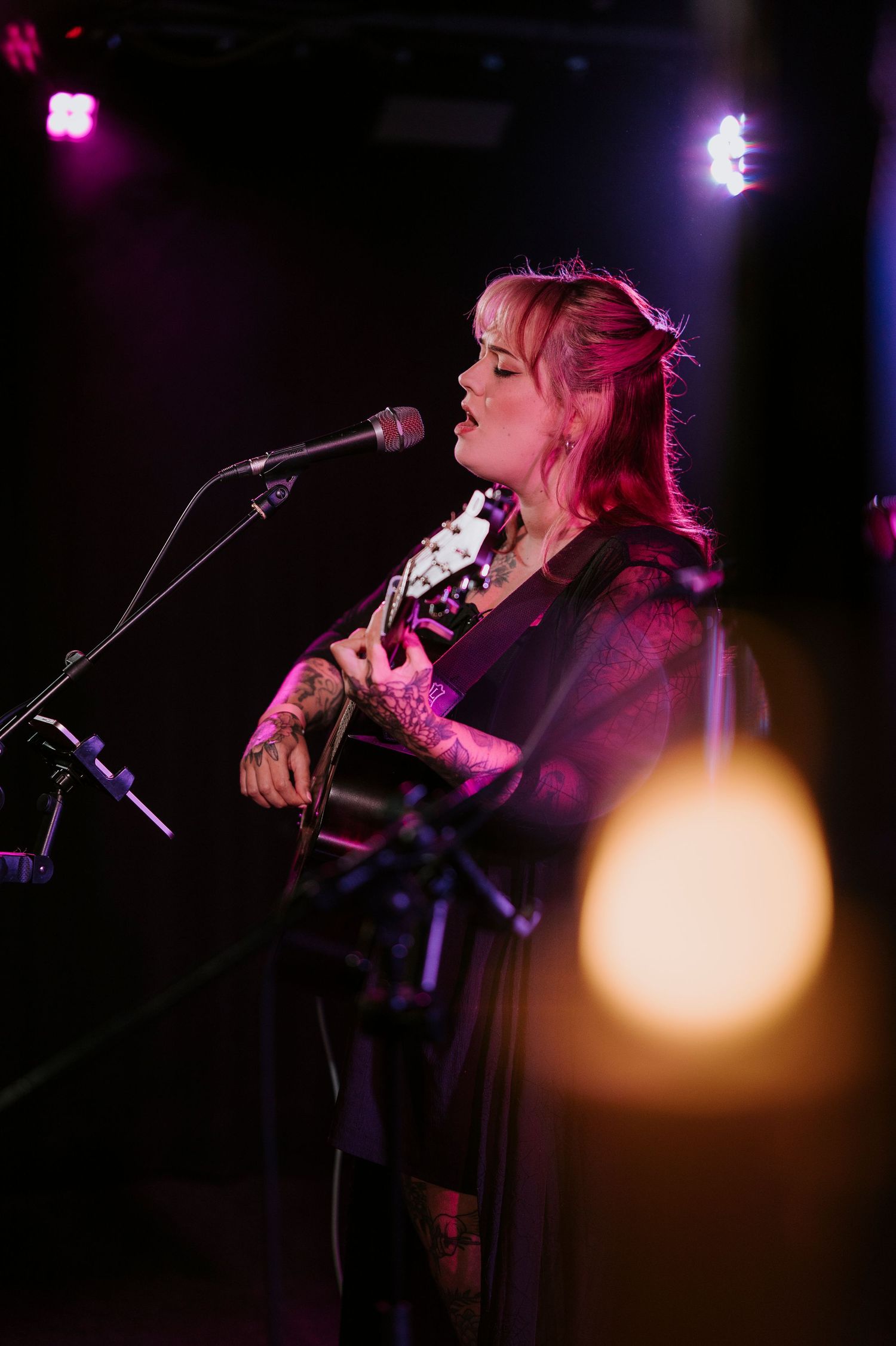 A performer sings into a microphone while playing acoustic guitar under dramatic purple stage lighting.