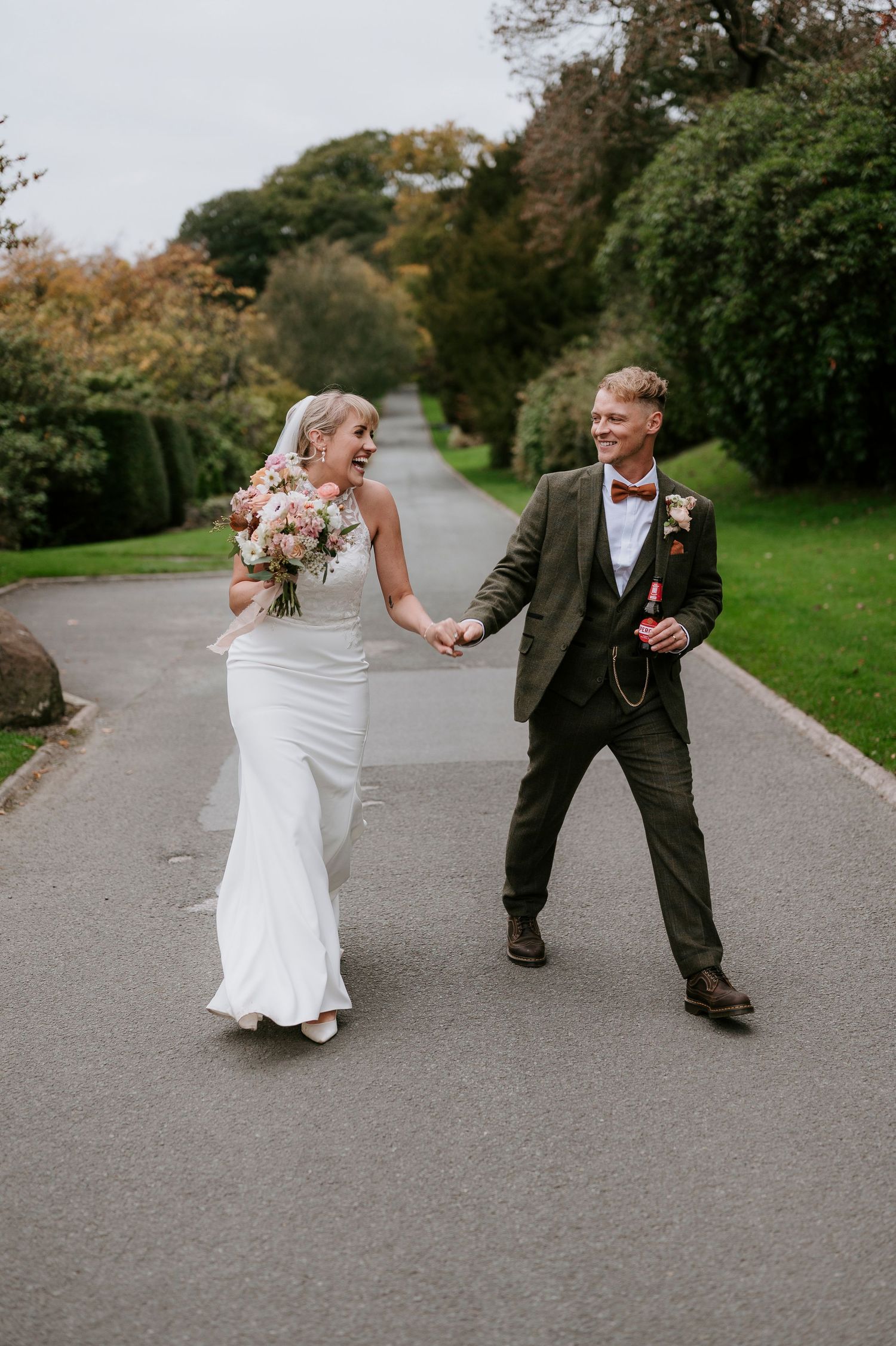 A couple in wedding attire dances playfully on a paved path surrounded by autumn greenery.