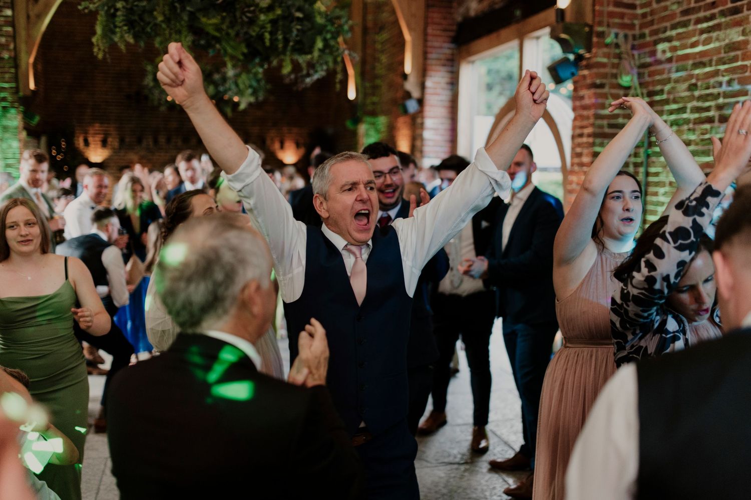Wedding guests dance and celebrate joyfully in a rustic brick-walled venue with string lights overhead.