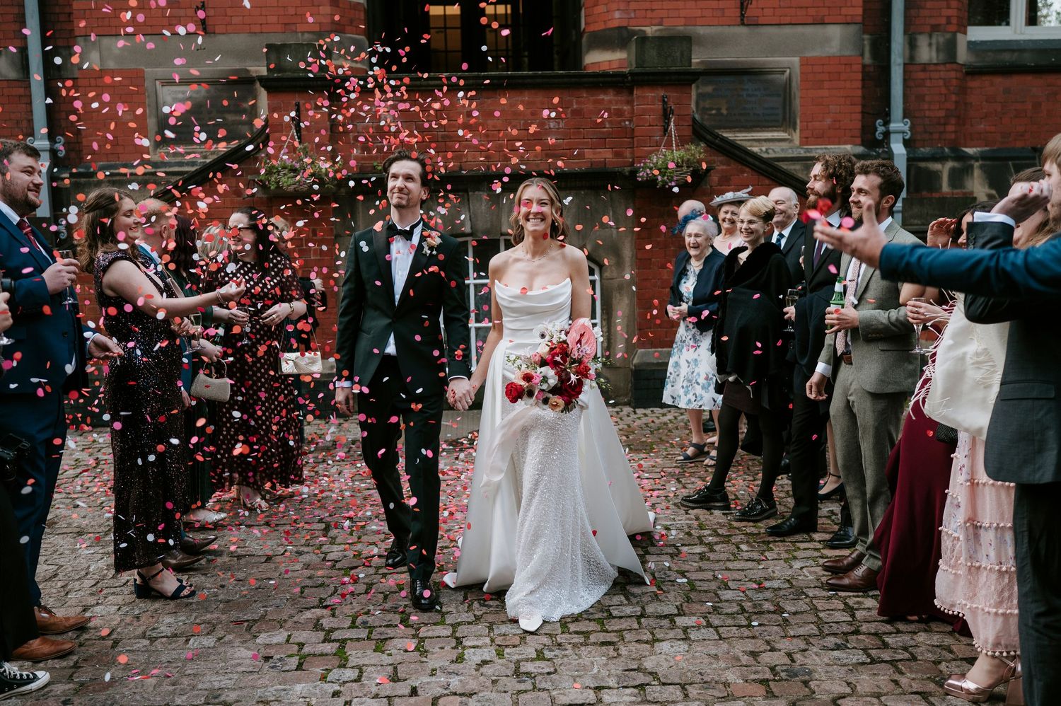 Wedding guests shower newlyweds with red confetti on cobblestone street outside historic brick building.