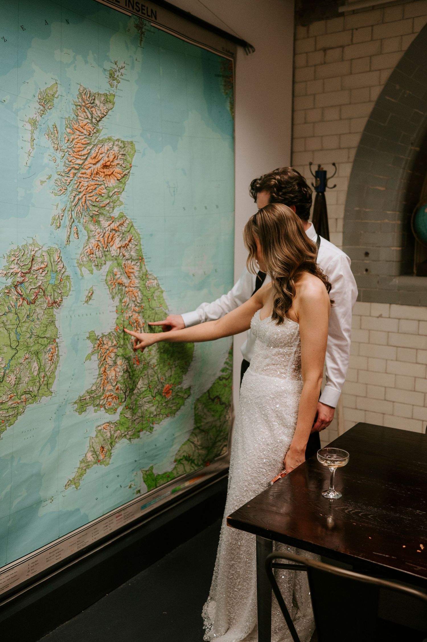 Someone in a white lace wedding dress points to a location on a vintage wall map of the British Isles.