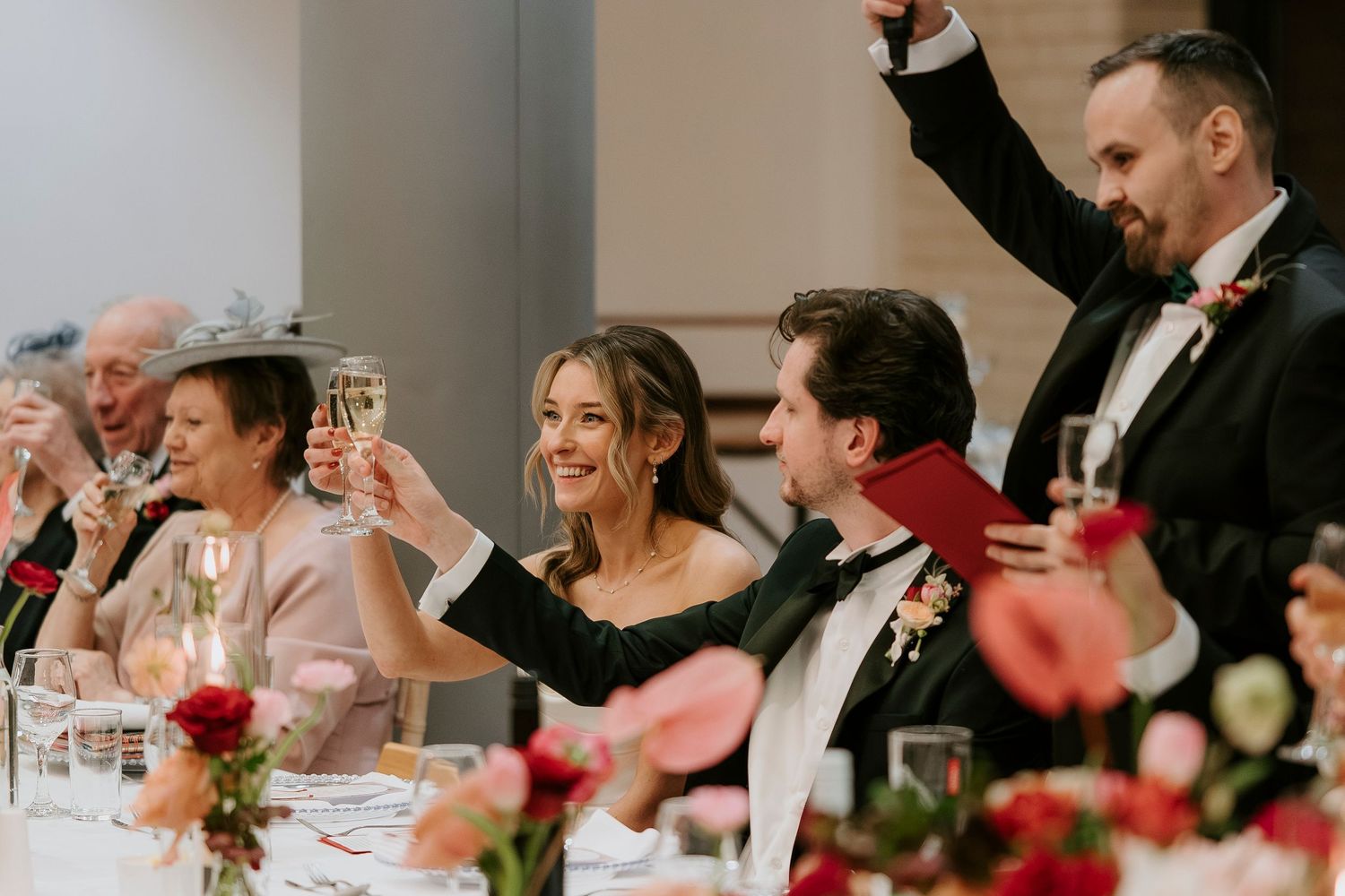 Wedding guests raise their glasses in a toast at an elegant reception with red floral centerpieces on the table.