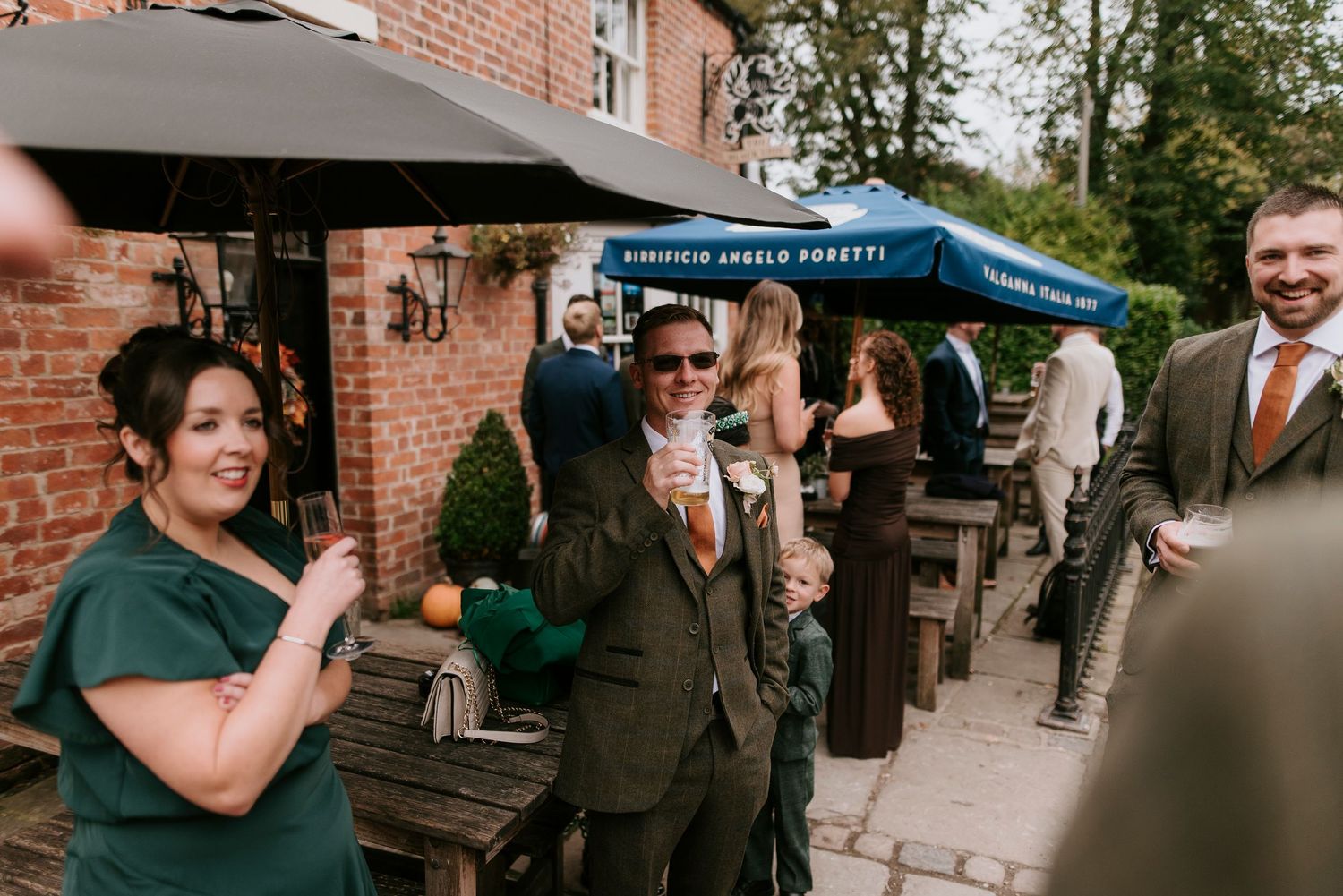 People in formal wear mingle and socialize at an outdoor wedding reception near a brick wall.