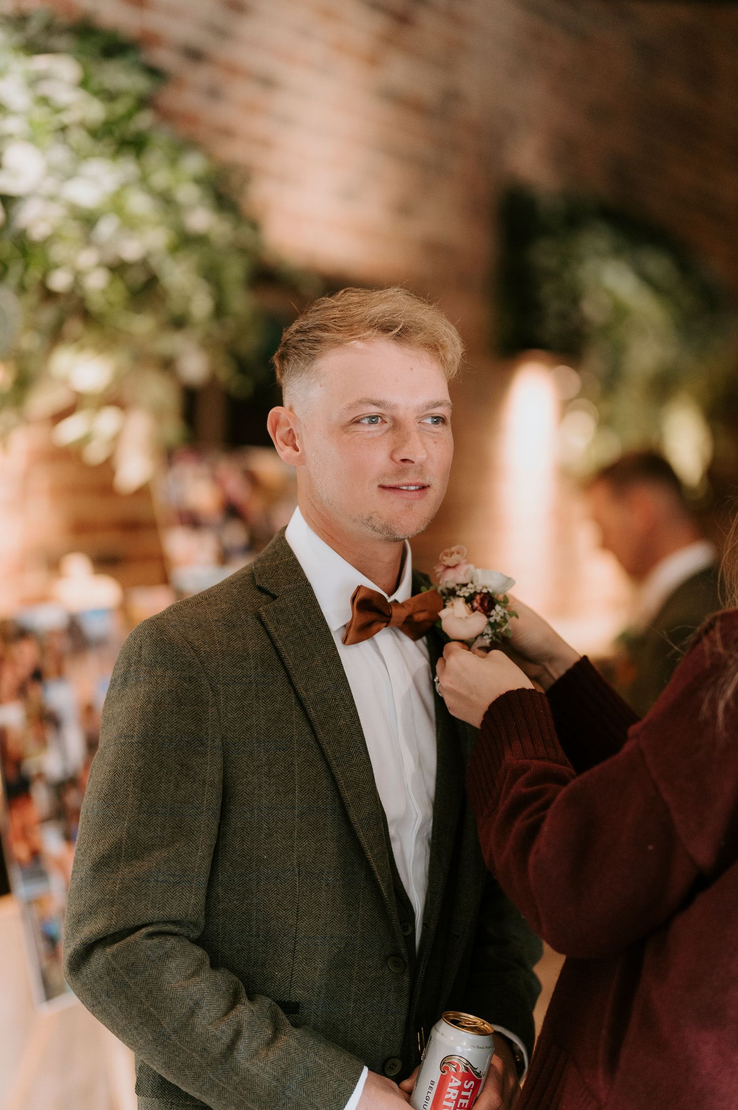 Person in formal attire and tweed jacket stands near brick wall at an event.