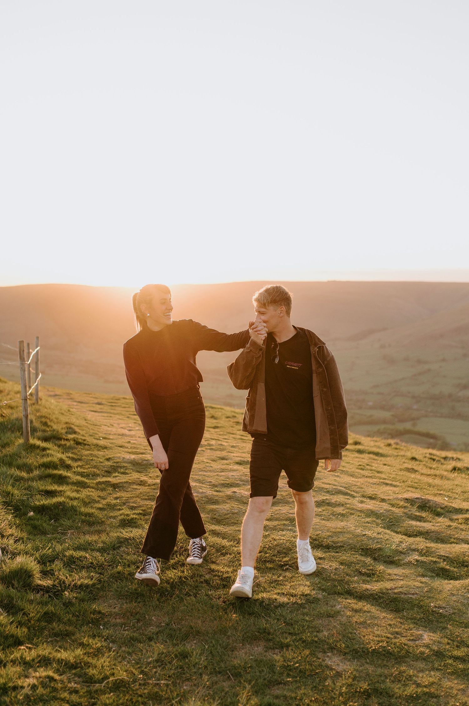 Two people walking together on a grassy hillside at sunset, silhouetted against golden rolling hills.