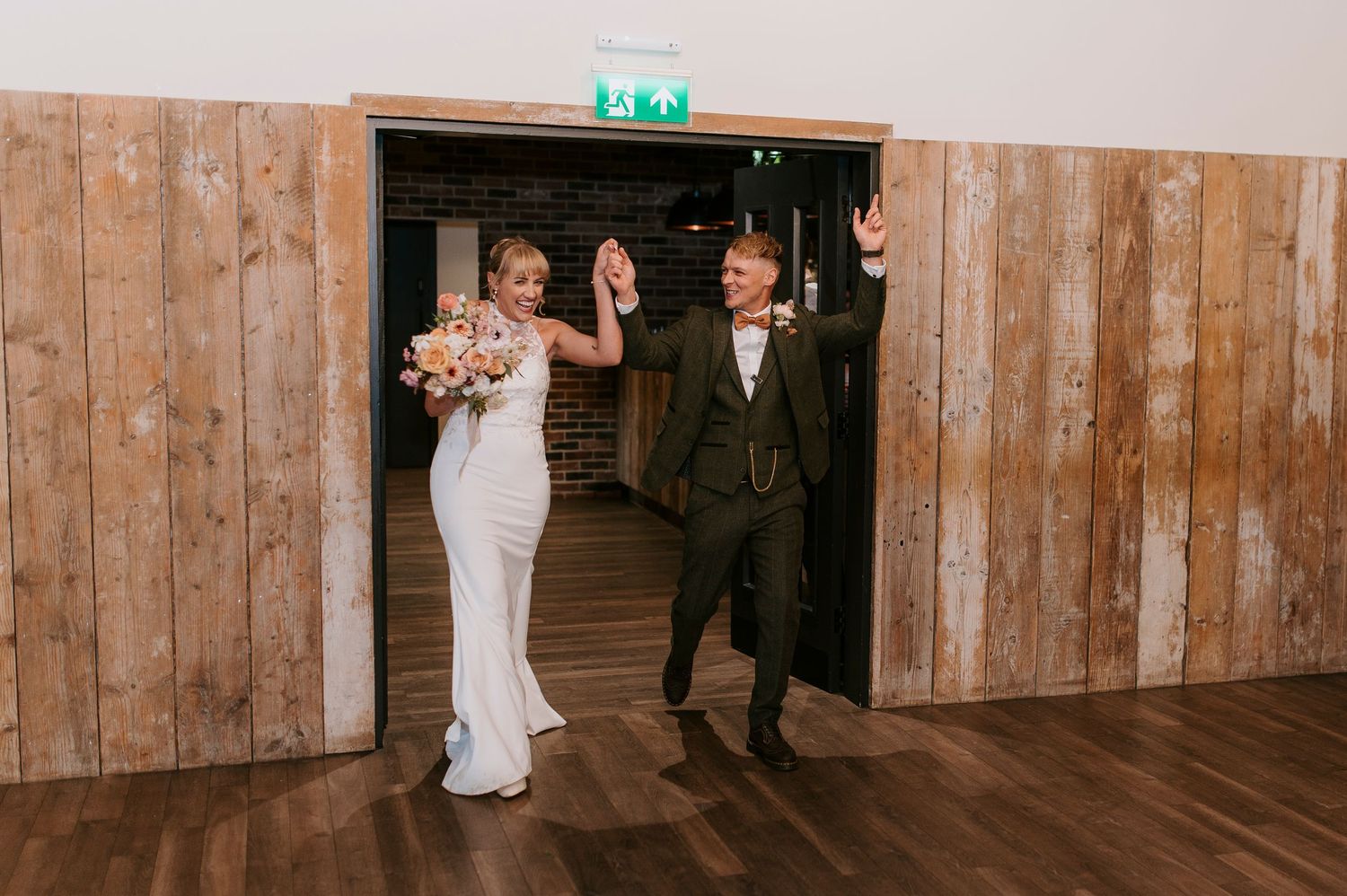 Newlyweds and wedding party walk down rustic wooden hallway in elegant formal attire.
