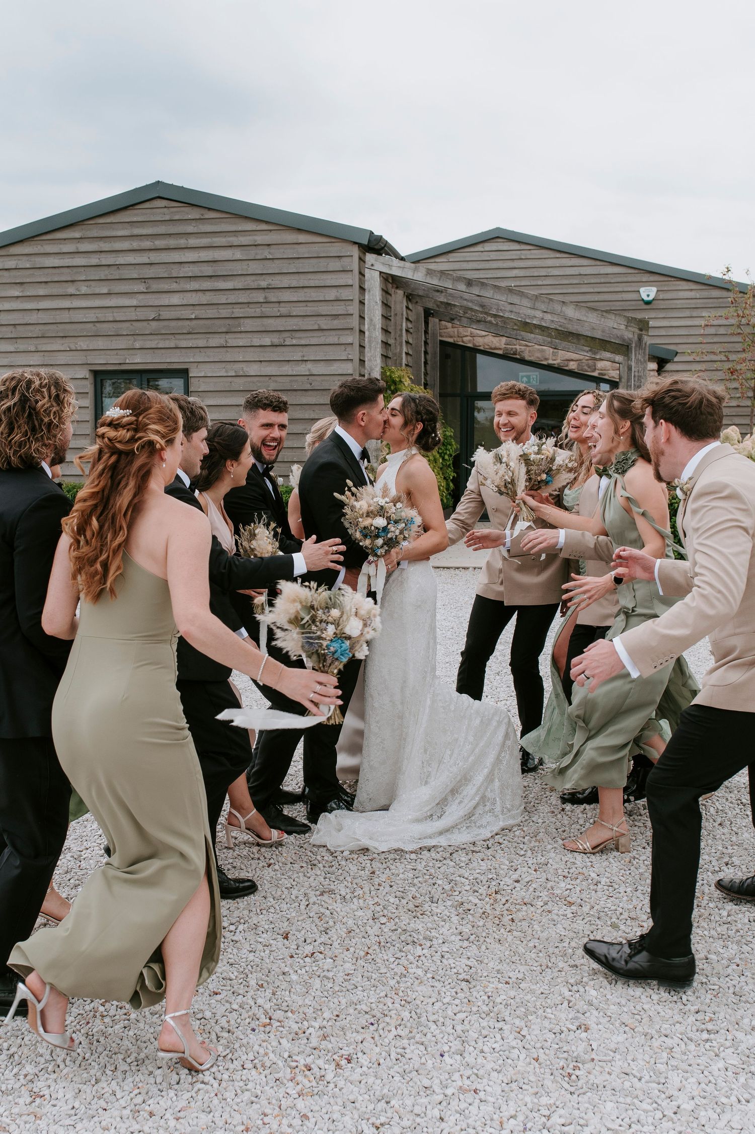 Wedding party celebrates outdoors on gravel with rustic wooden buildings in background.