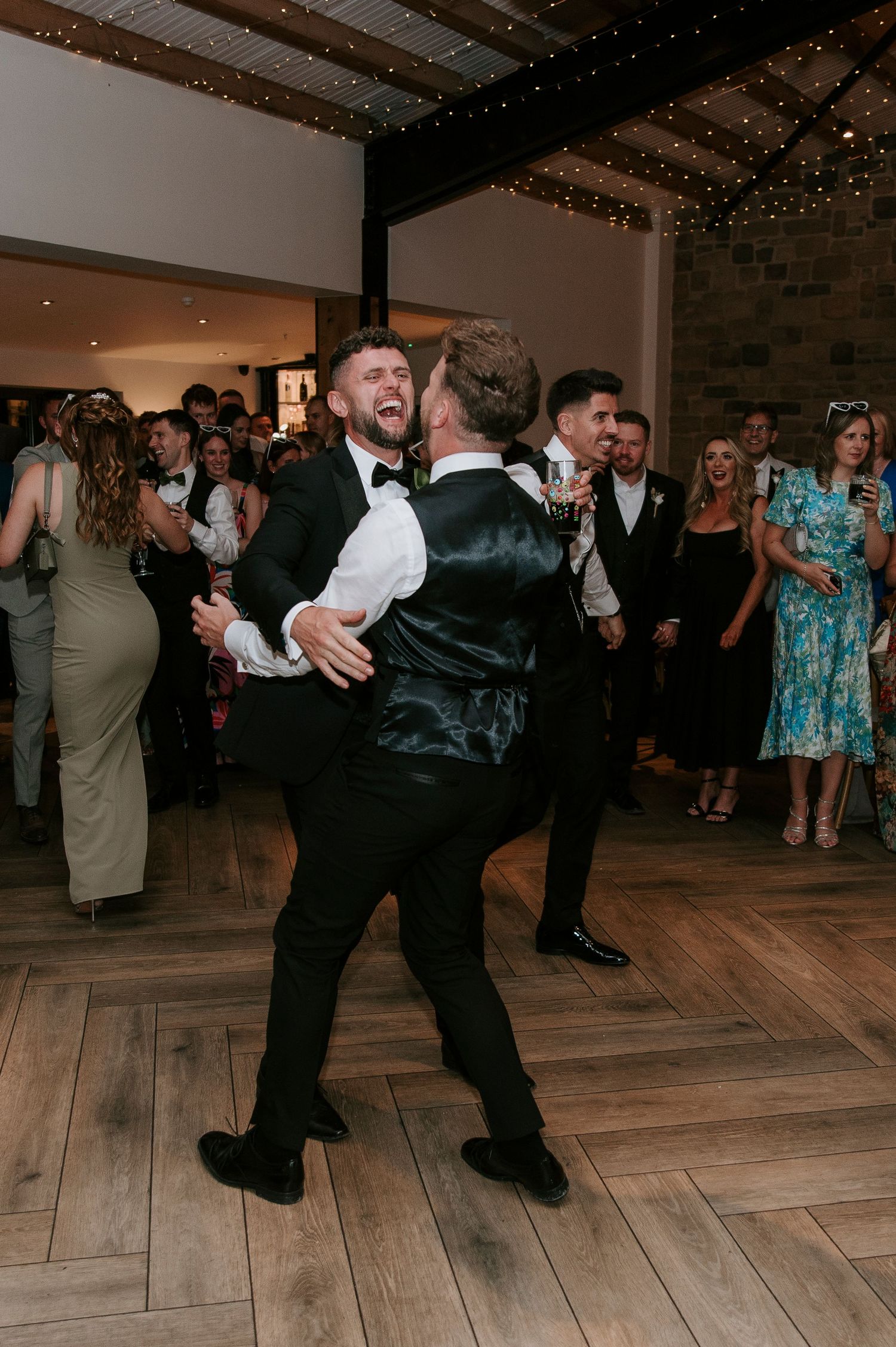 Two wedding guests perform a choreographed dance routine on a wooden floor at a reception celebration.