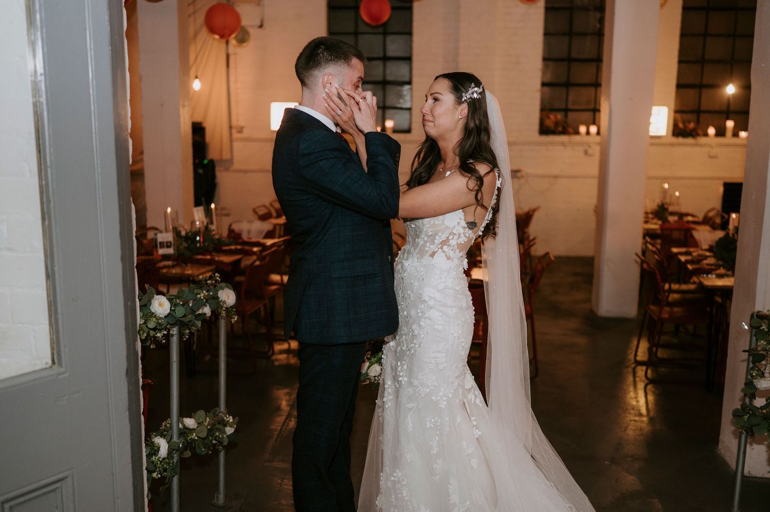 Bride tenderly holds groom's face during first dance at elegant indoor wedding reception with candlelit tables.