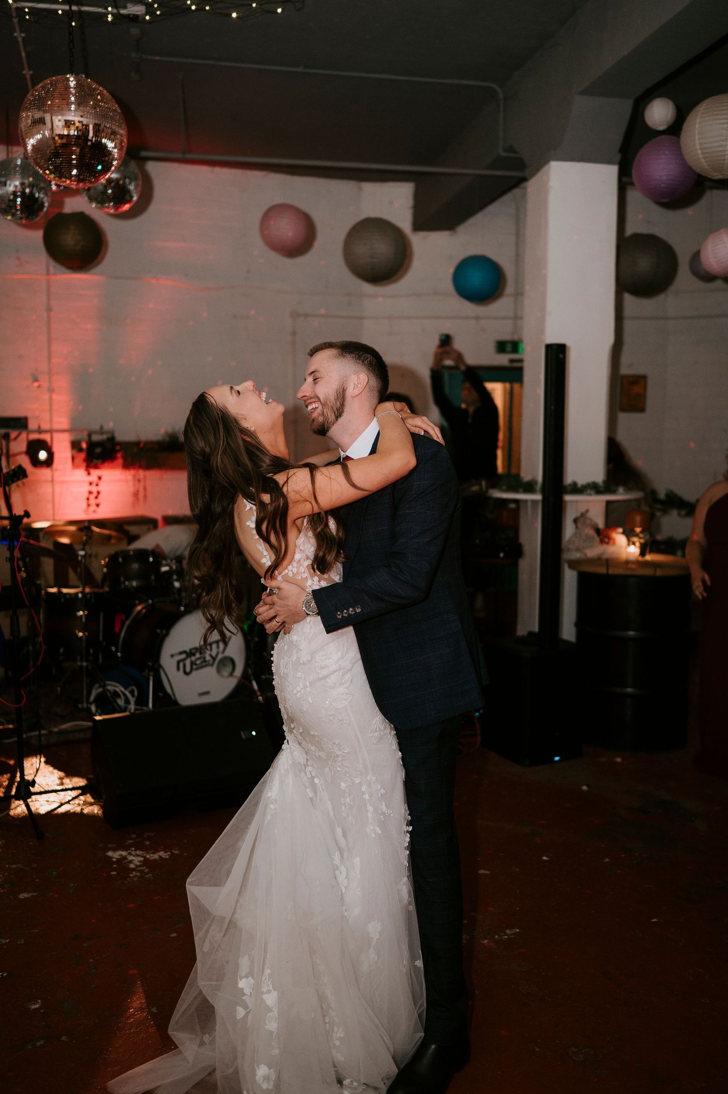 Bride in lace gown and groom share first dance under colorful paper lanterns at wedding reception.