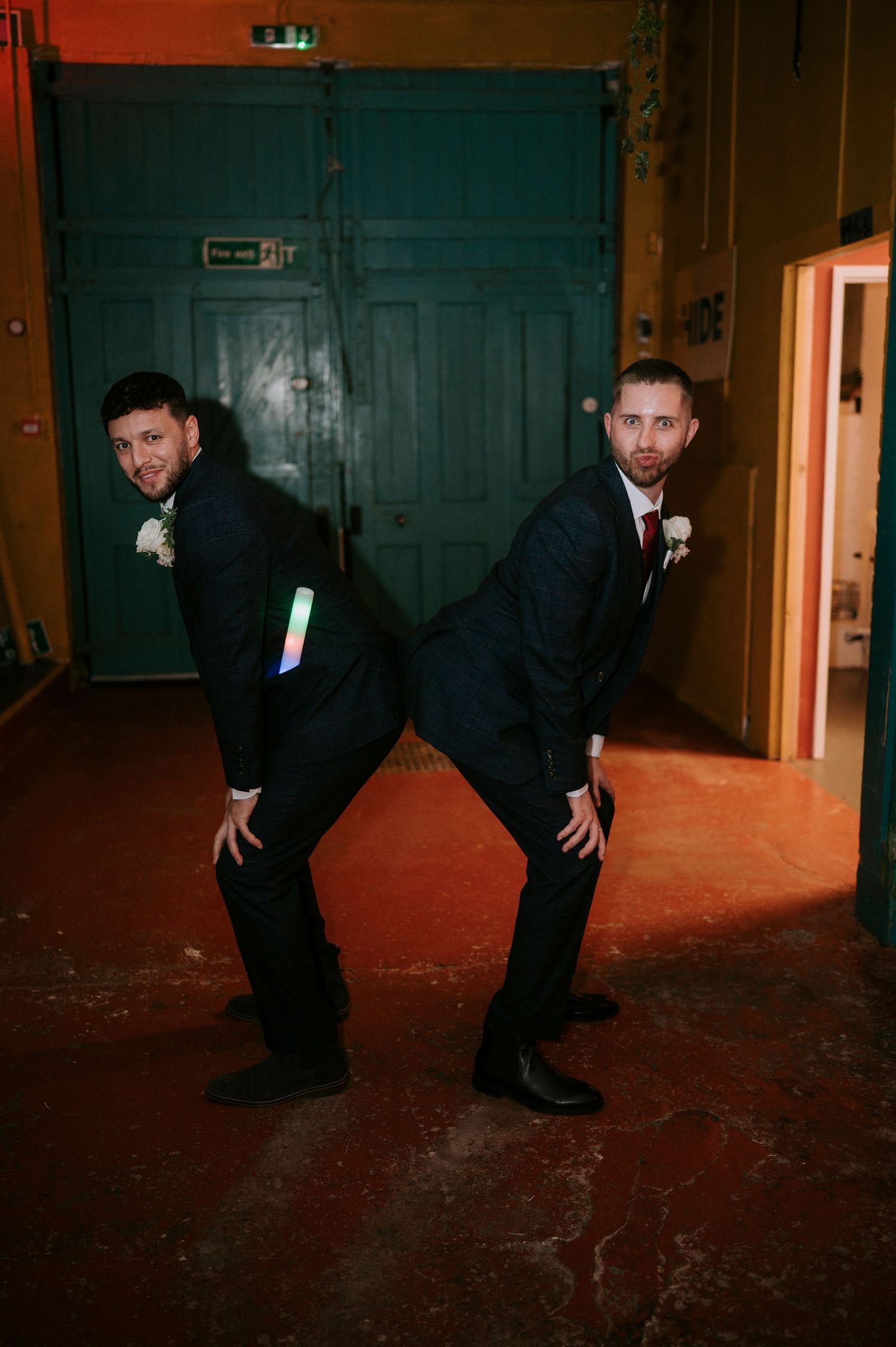 Two groomsmen in black suits pose back-to-back with glow sticks at a wedding reception.