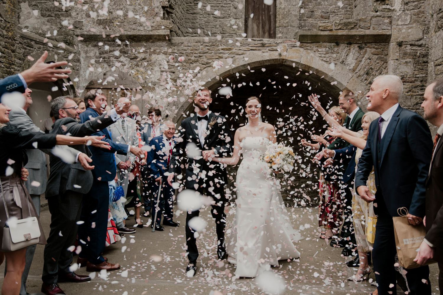 Wedding celebration with guests throwing confetti at couple outside historic brick building.