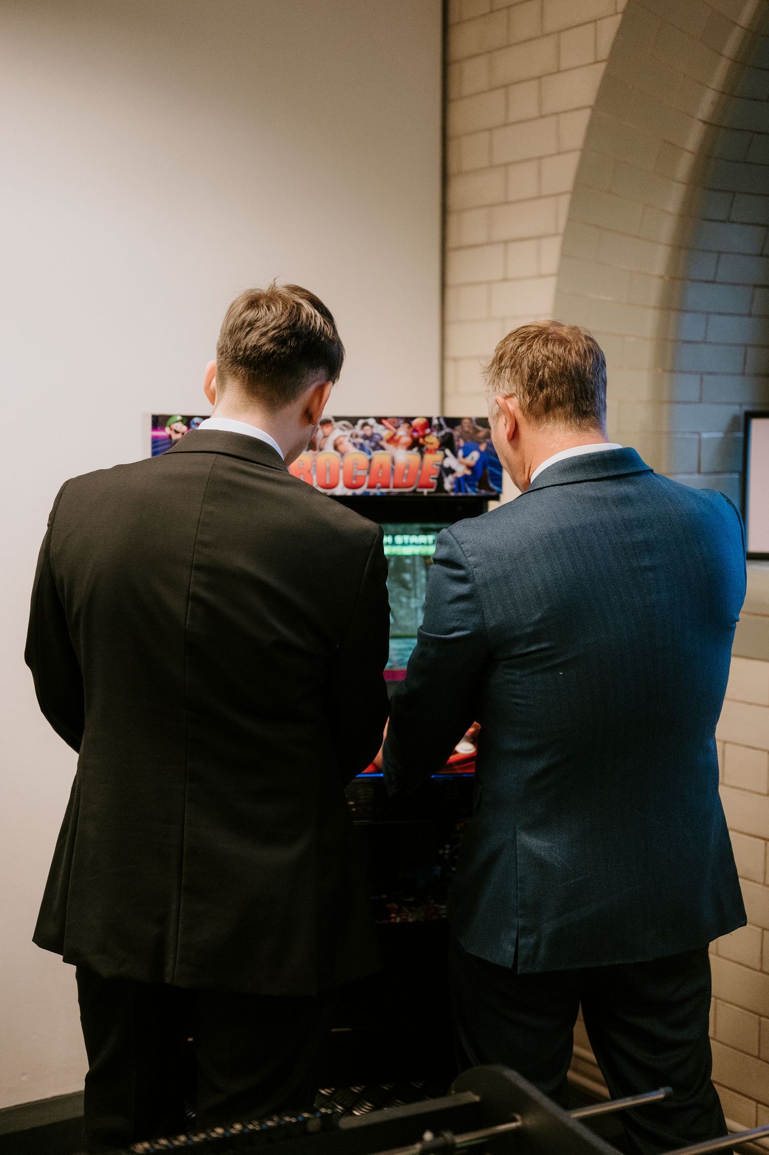 Two people in suits sit side by side in front of a screen in a dimly lit room.