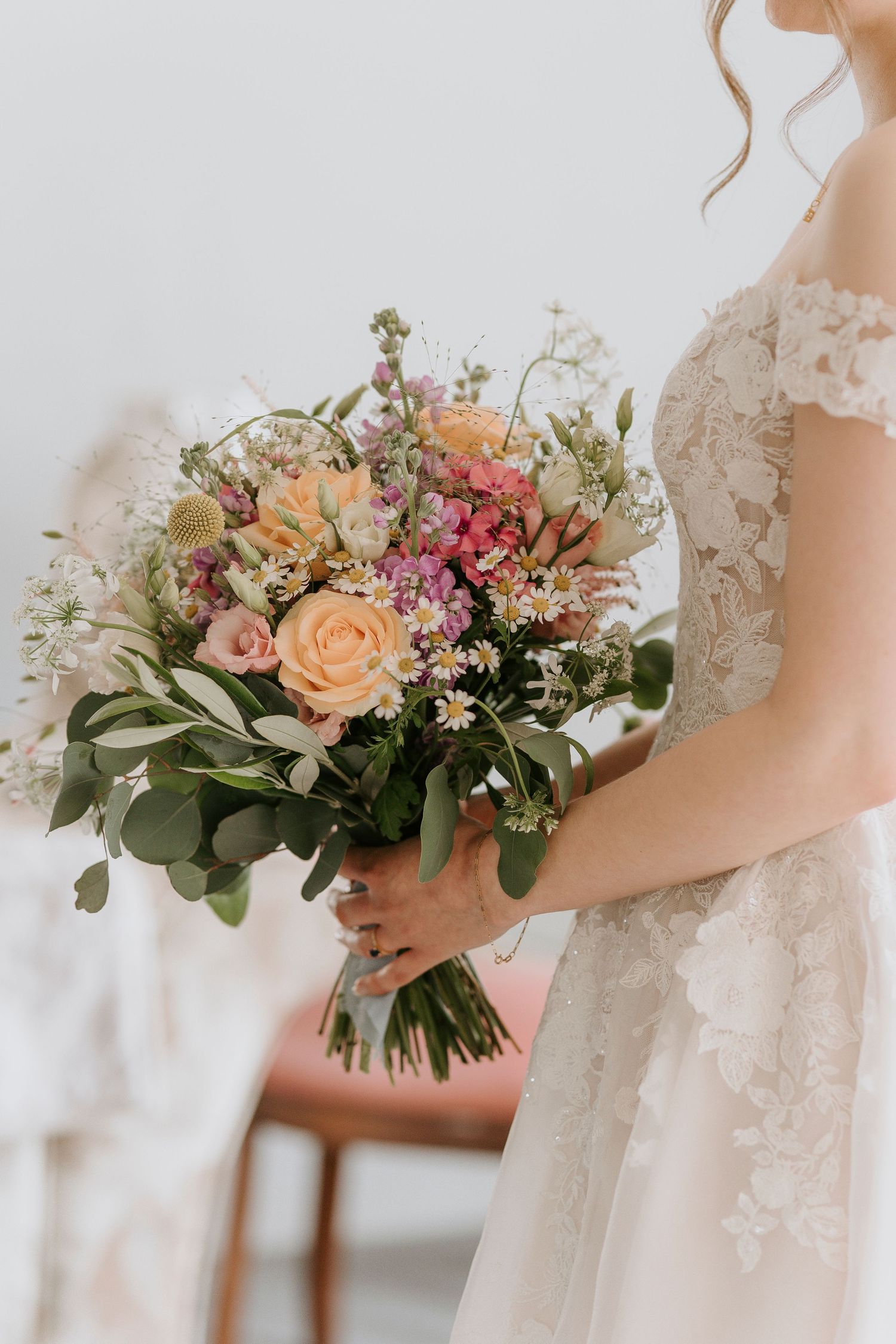 A delicate bridal bouquet with peach roses, pink flowers and eucalyptus leaves held against a white lace wedding dress.