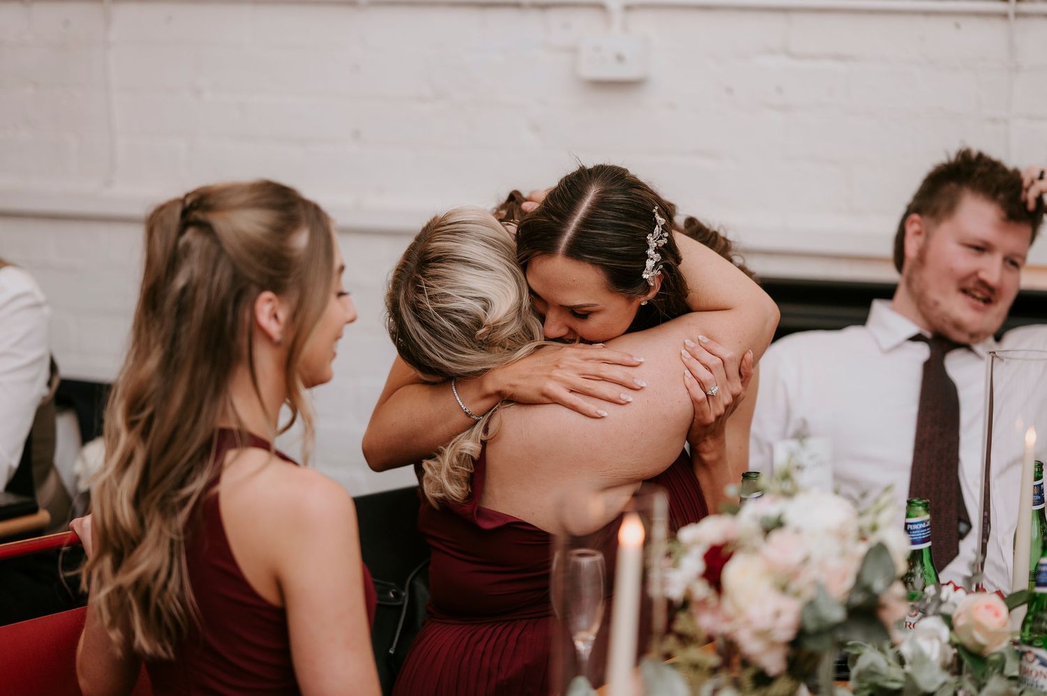 Bride receives warm embrace from bridesmaid in burgundy dress at wedding reception dinner table.