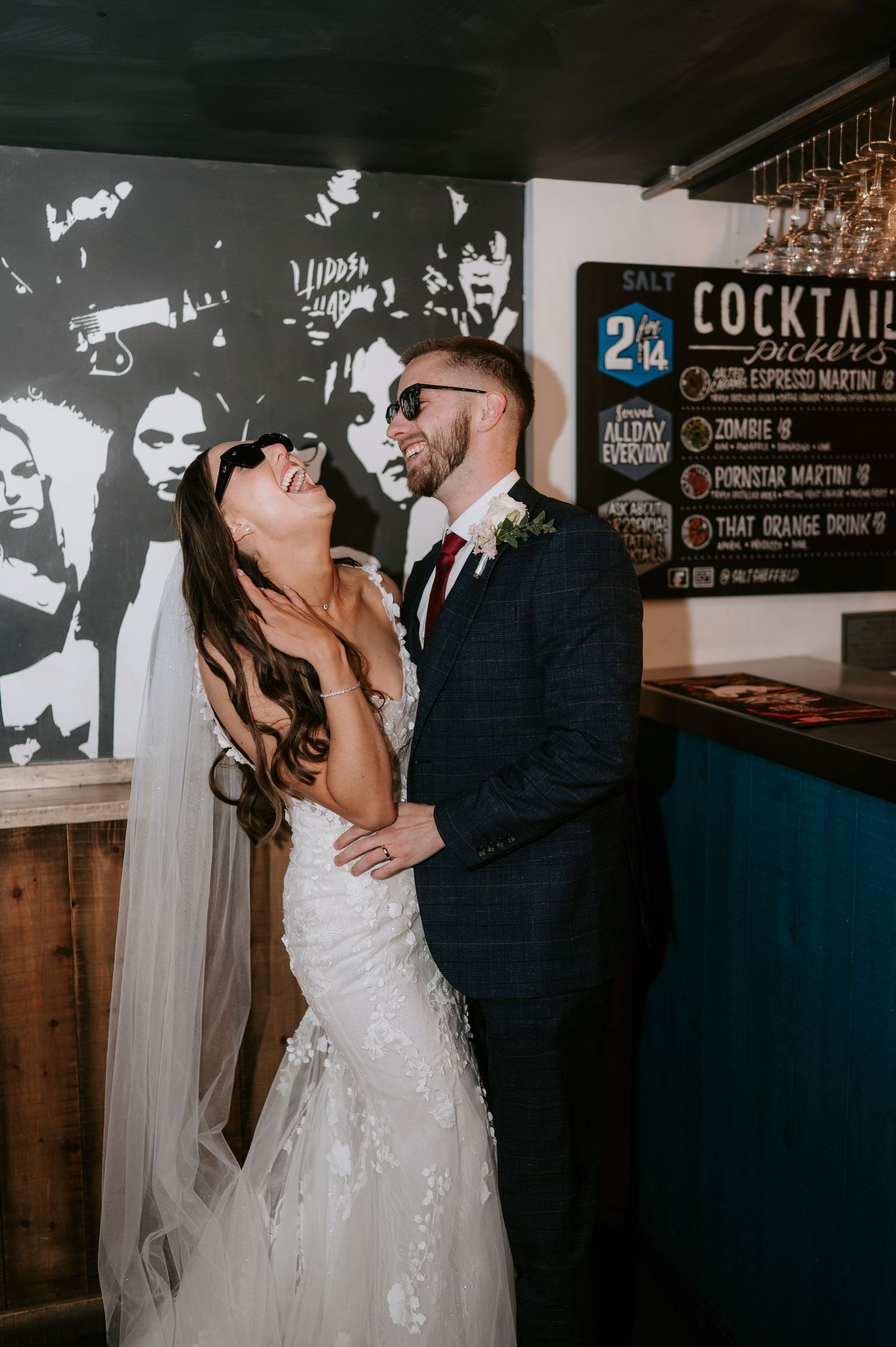 Laughing newlyweds wearing sunglasses share a fun moment at a cocktail bar on their wedding day.
