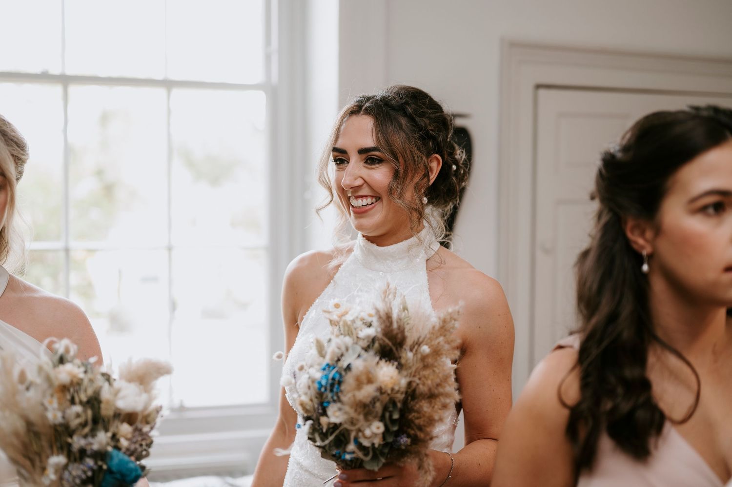 Bridesmaids in elegant white halter dresses hold delicate bouquets of dried flowers while smiling by a bright window.