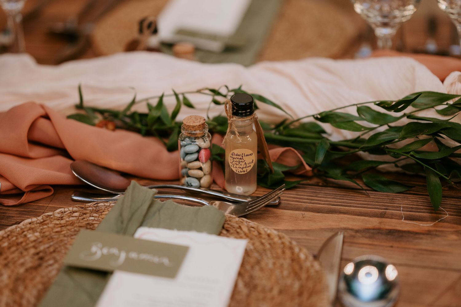 Place setting with woven placemats, green napkins, tropical leaves and small glass bottles on a rustic wooden table.