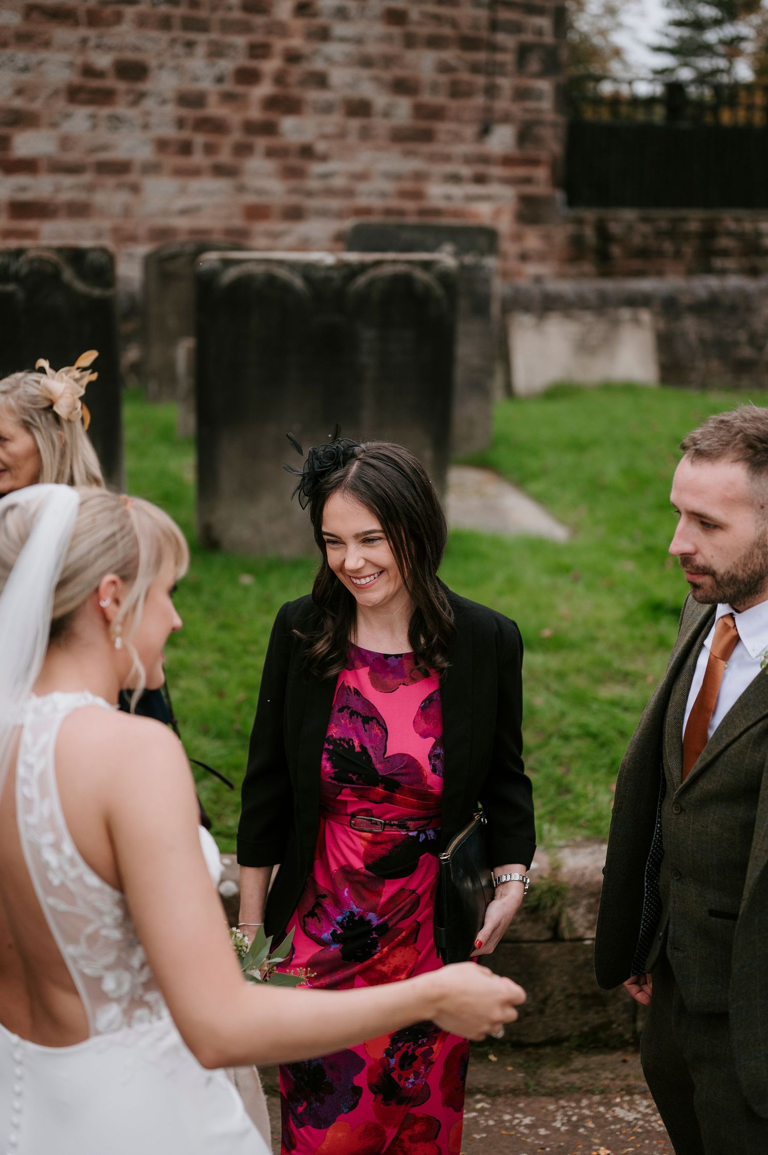 Wedding party members share laughter and conversation outside a rustic brick building during outdoor celebration.