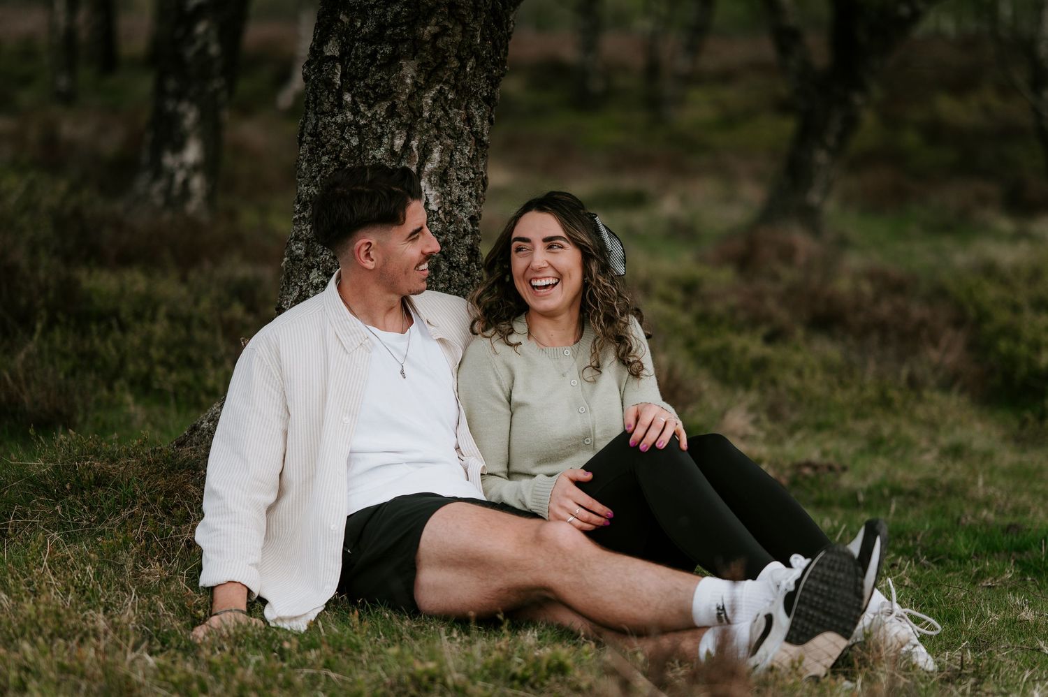 A couple sits together on grassy terrain during a romantic outdoor photoshoot at golden hour.