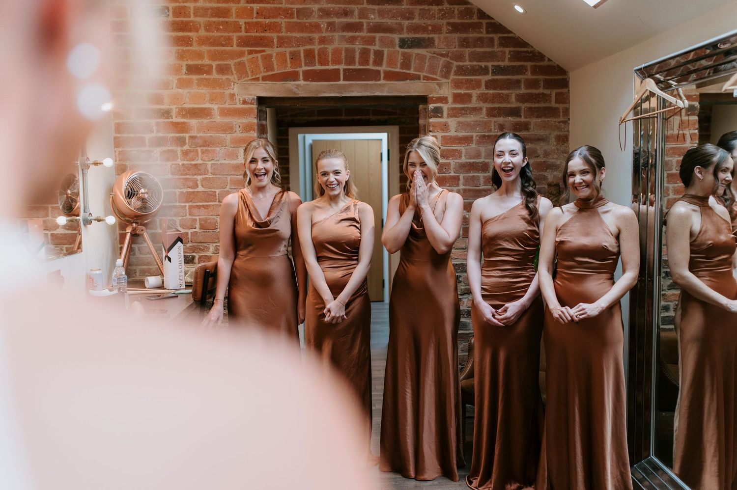 A group wearing matching rust-colored bridesmaid dresses stands together in a brick-walled room.