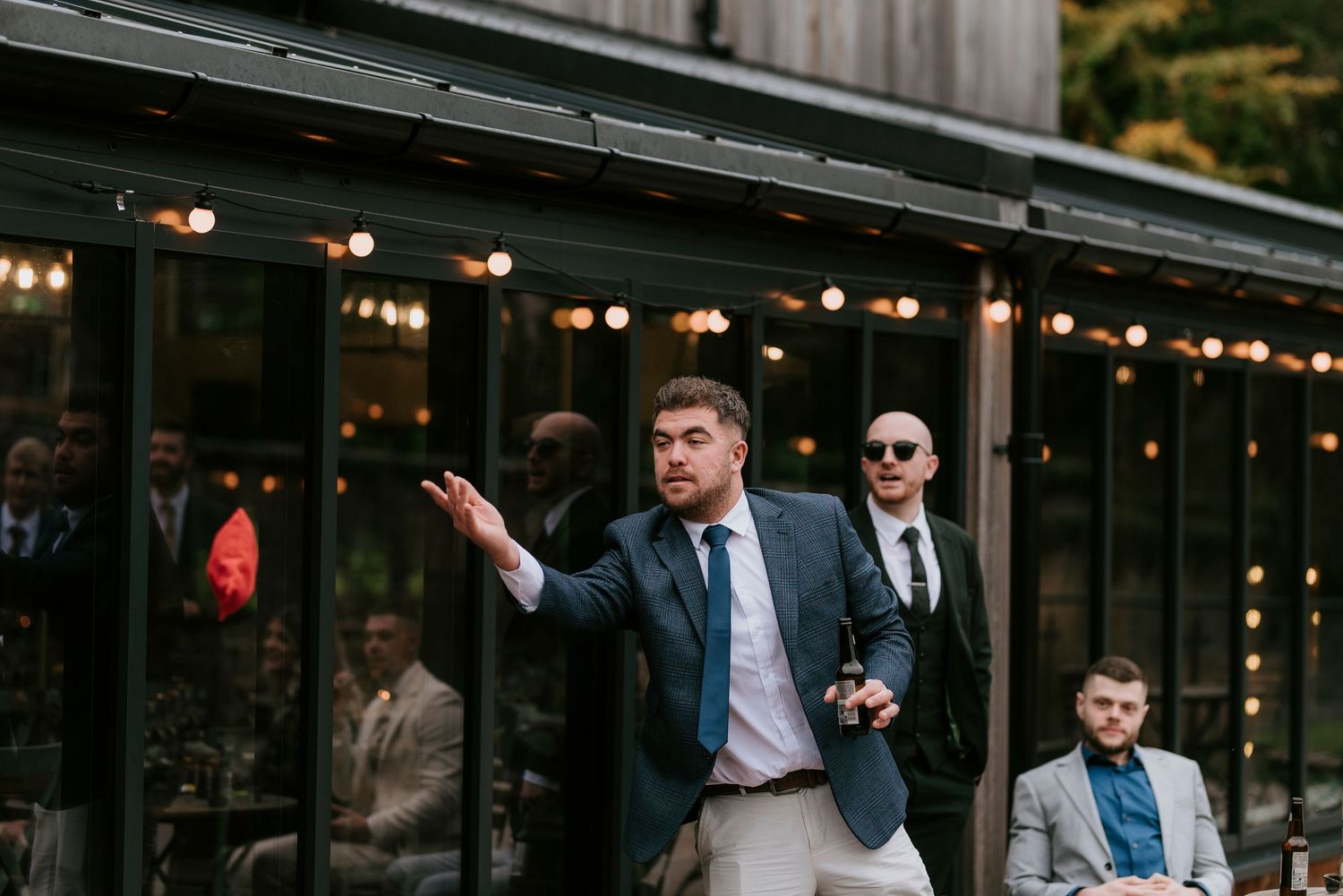Wedding guests gather outside a rustic venue decorated with string lights at dusk.