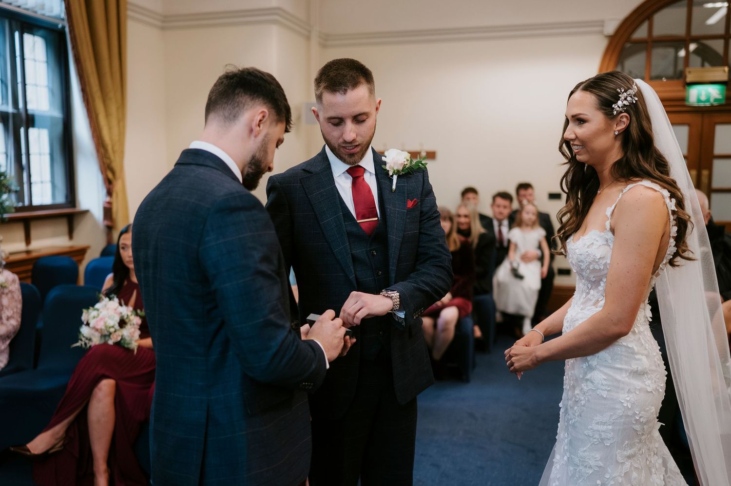 Groom exchanges wedding rings with best man as bride watches during an elegant indoor wedding ceremony.