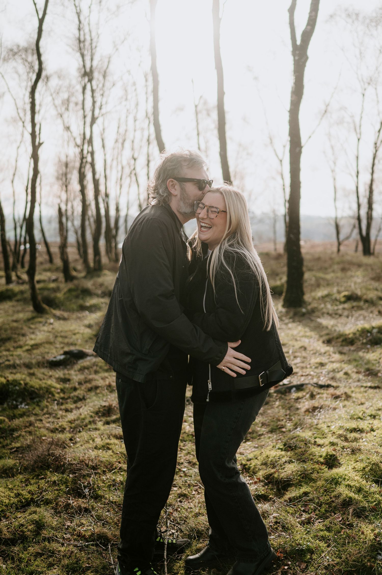 A couple in black outfits shares intimate moments while walking through a misty forest with sunlight filtering through trees.