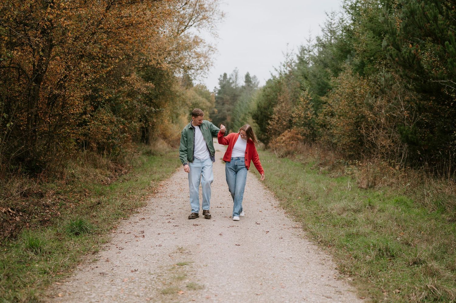 A couple walks along a scenic autumn trail surrounded by trees and fallen leaves.