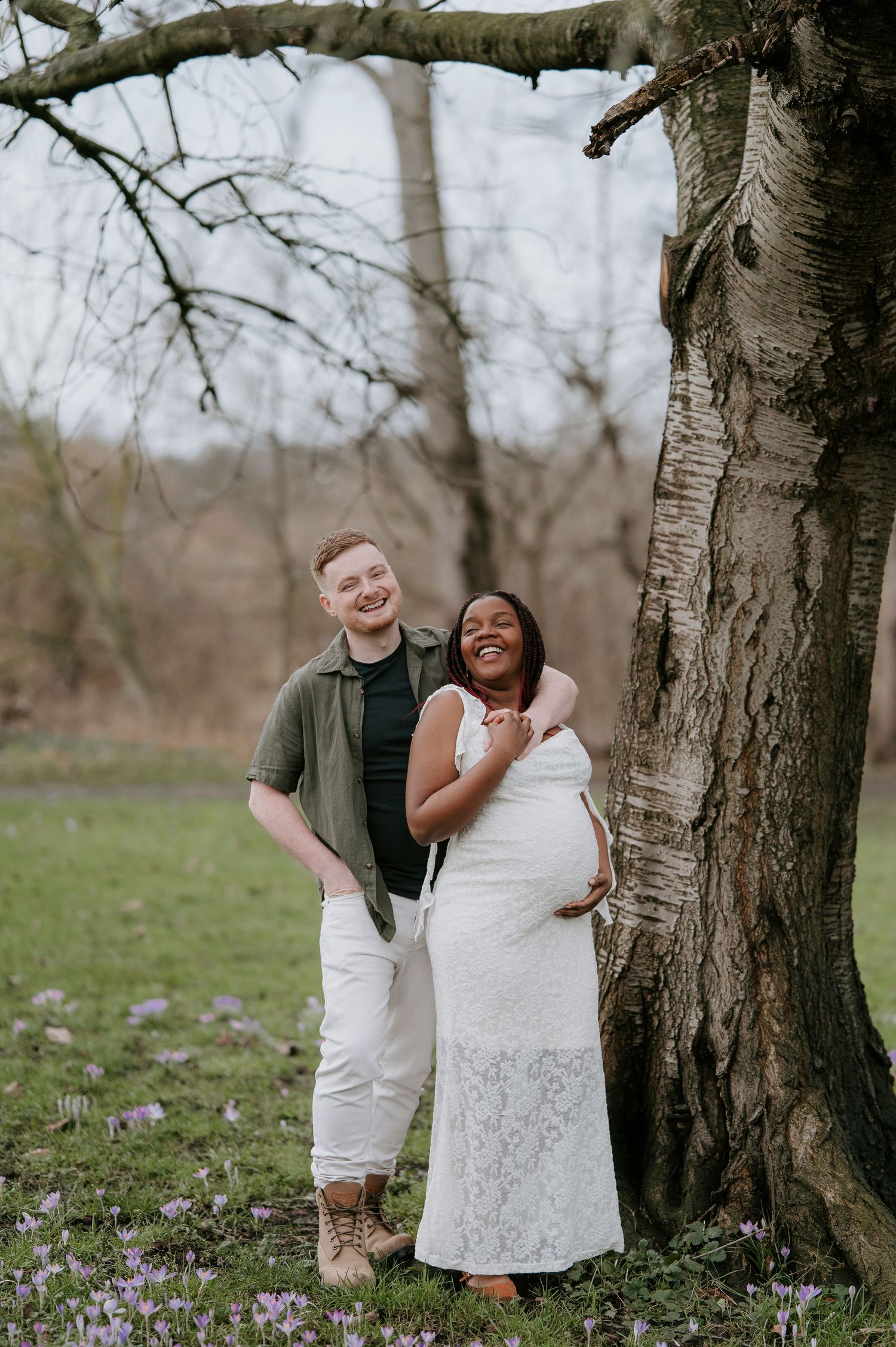 A couple shares a joyful moment together under a large tree with purple wildflowers blooming in the grass.