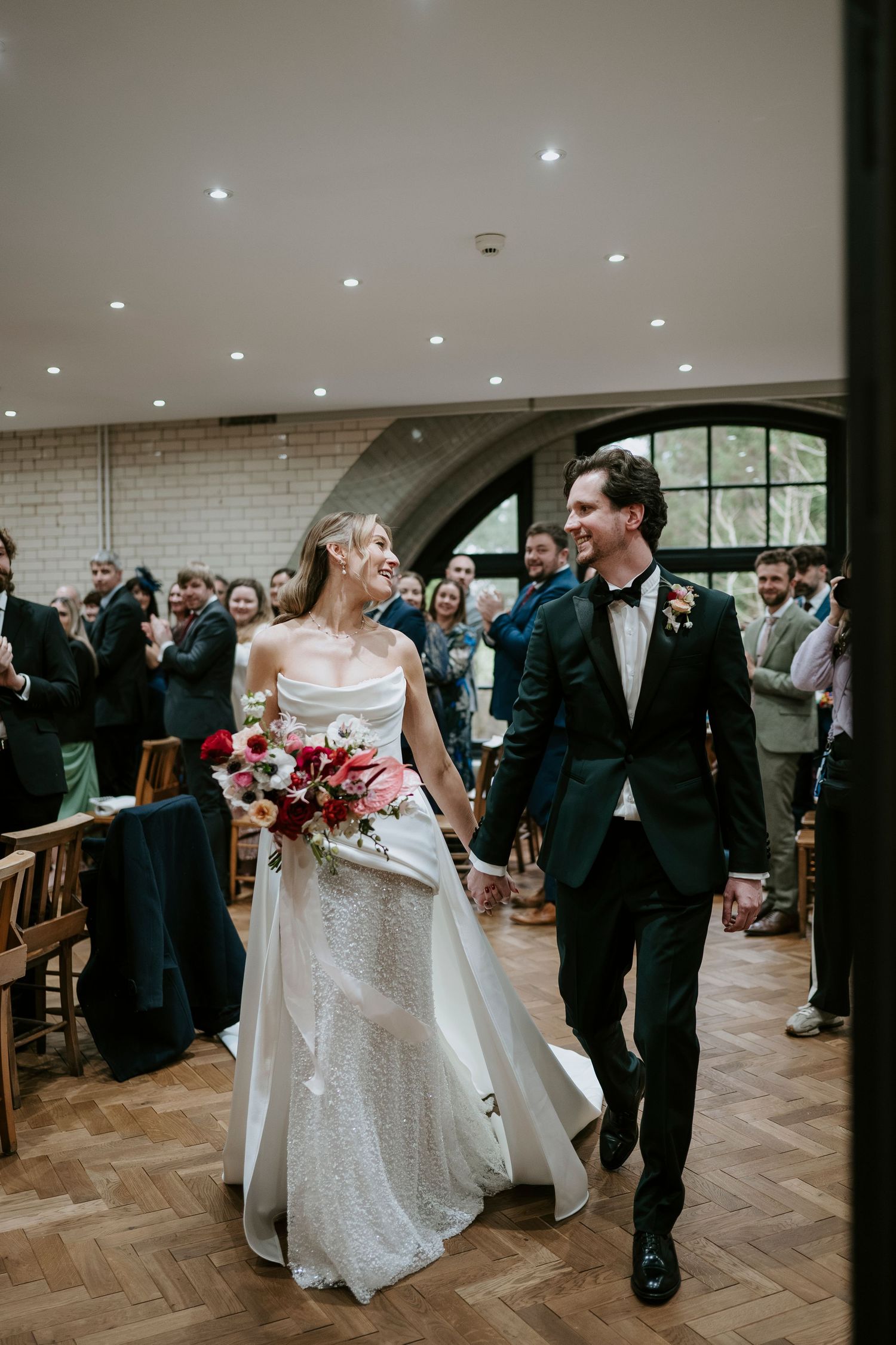 Newlyweds dance together in a bright reception hall with herringbone floors and white brick walls.