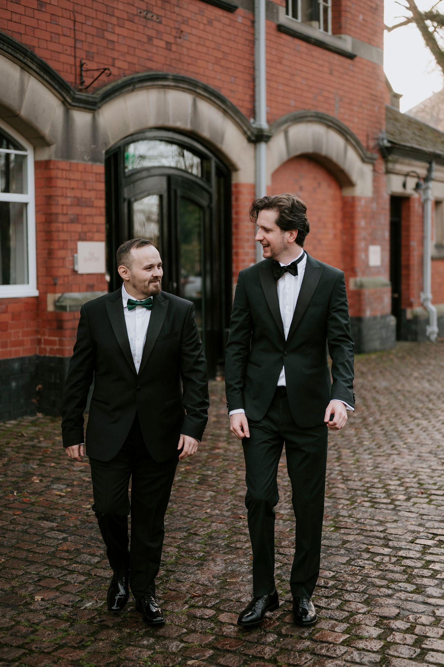 Groomsmen in black tuxedos share laughs while walking along a cobblestone path by a brick building.
