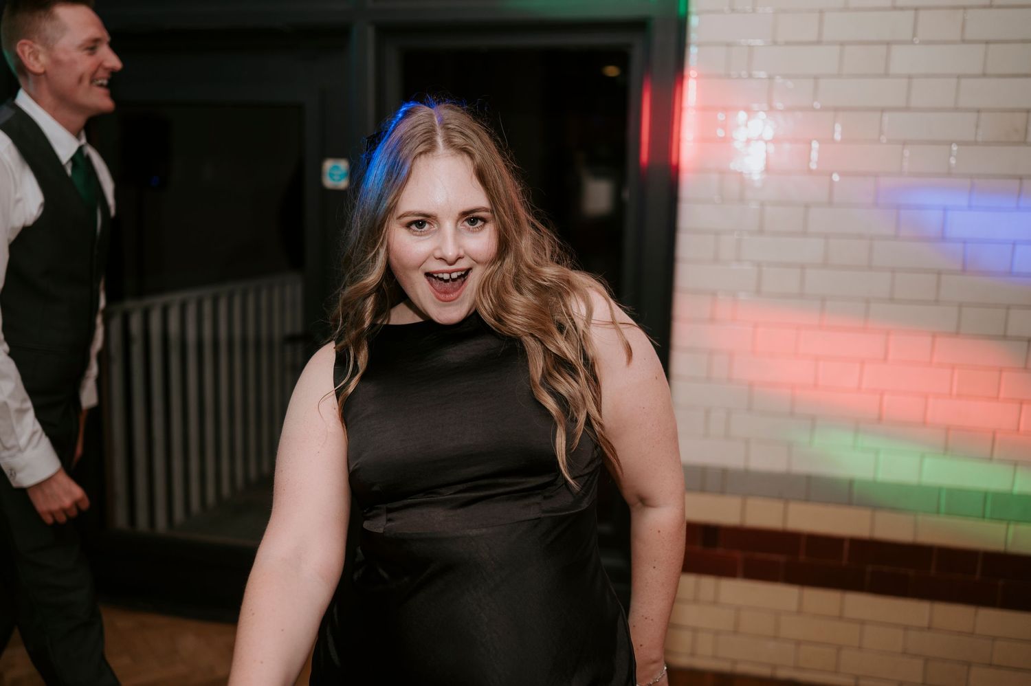 A person in a black formal dress poses at a party with colorful lighting in the background.