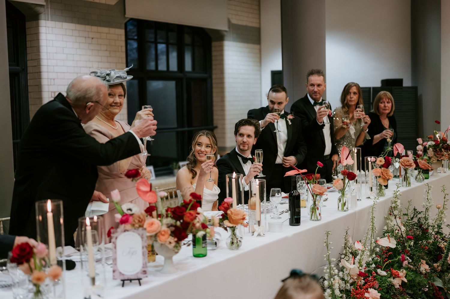 Wedding party laughs together at long reception table decorated with floral arrangements and glowing candles.