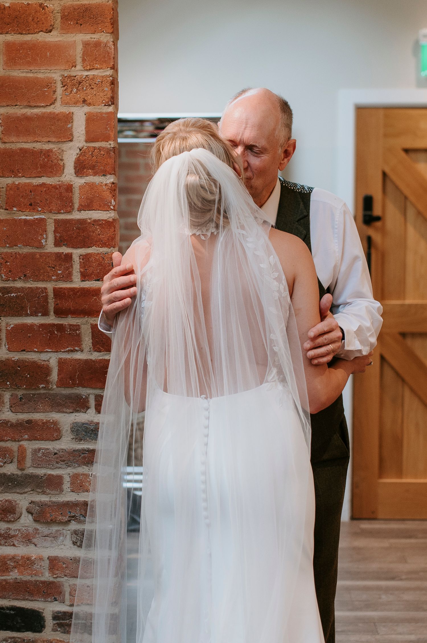 Emotional first look moment between bride in flowing veil and father at indoor brick-walled venue.