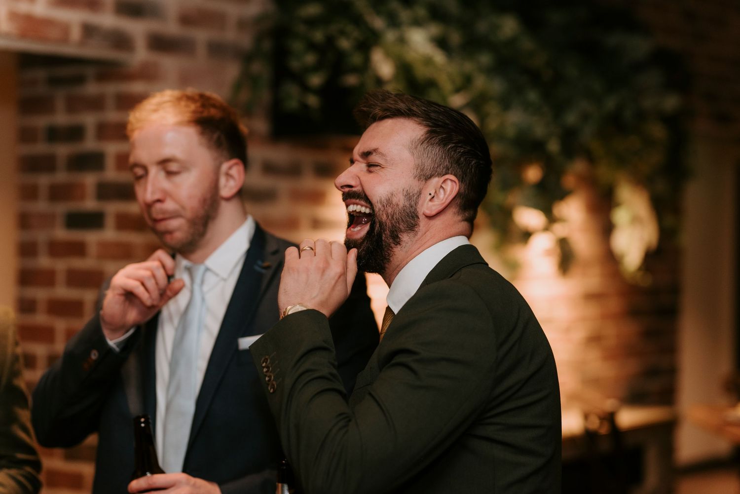 Group of people in formal attire laughing and enjoying themselves at an elegant brick-walled venue.