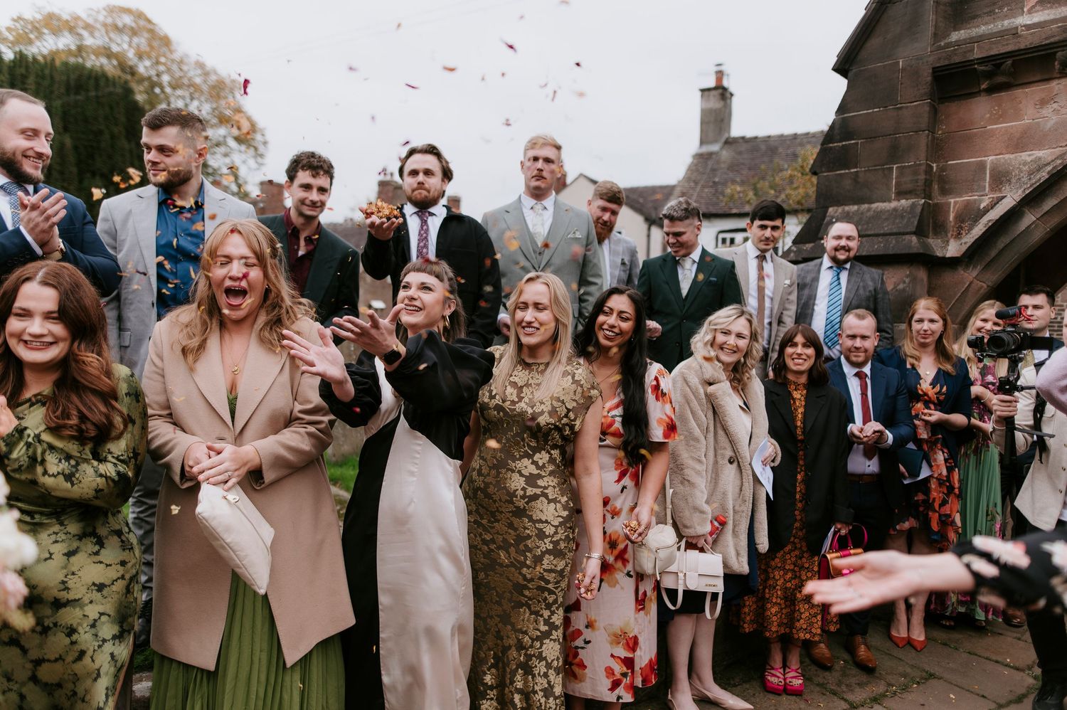 Wedding guests stand together outside a rustic stone church building during a celebration.