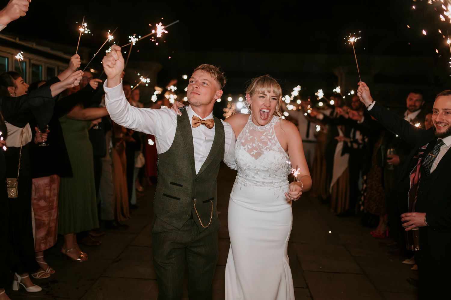 Wedding celebration at night with sparklers illuminating guests as they cheer and wave their arms in celebration.