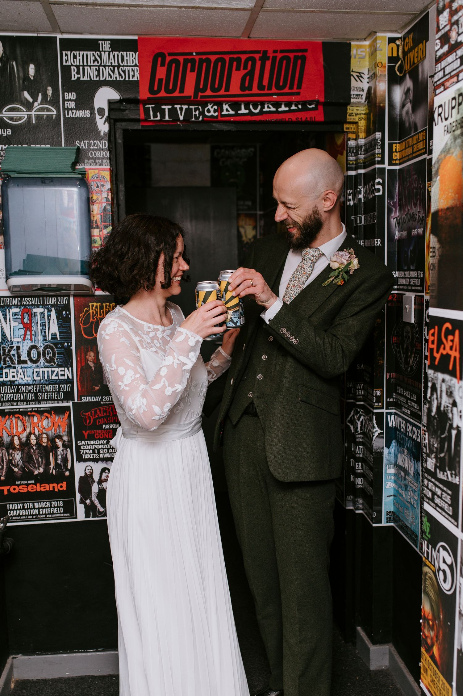 A couple in wedding attire shares a drink in a record store aisle surrounded by posters and album covers.