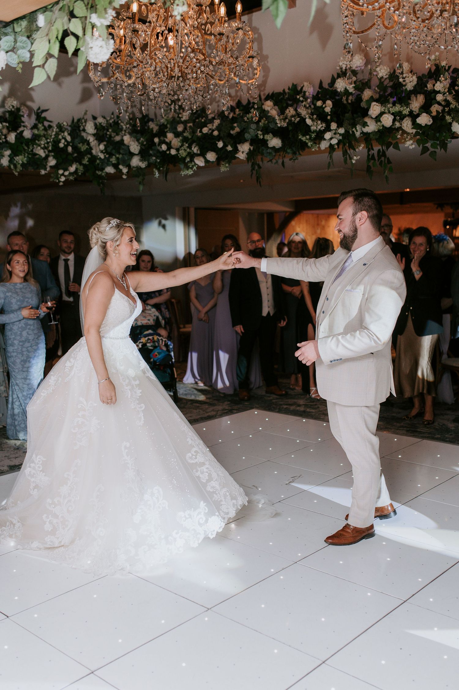 Couple shares their first dance at wedding reception under crystal chandeliers and floral decorations on white dance floor.