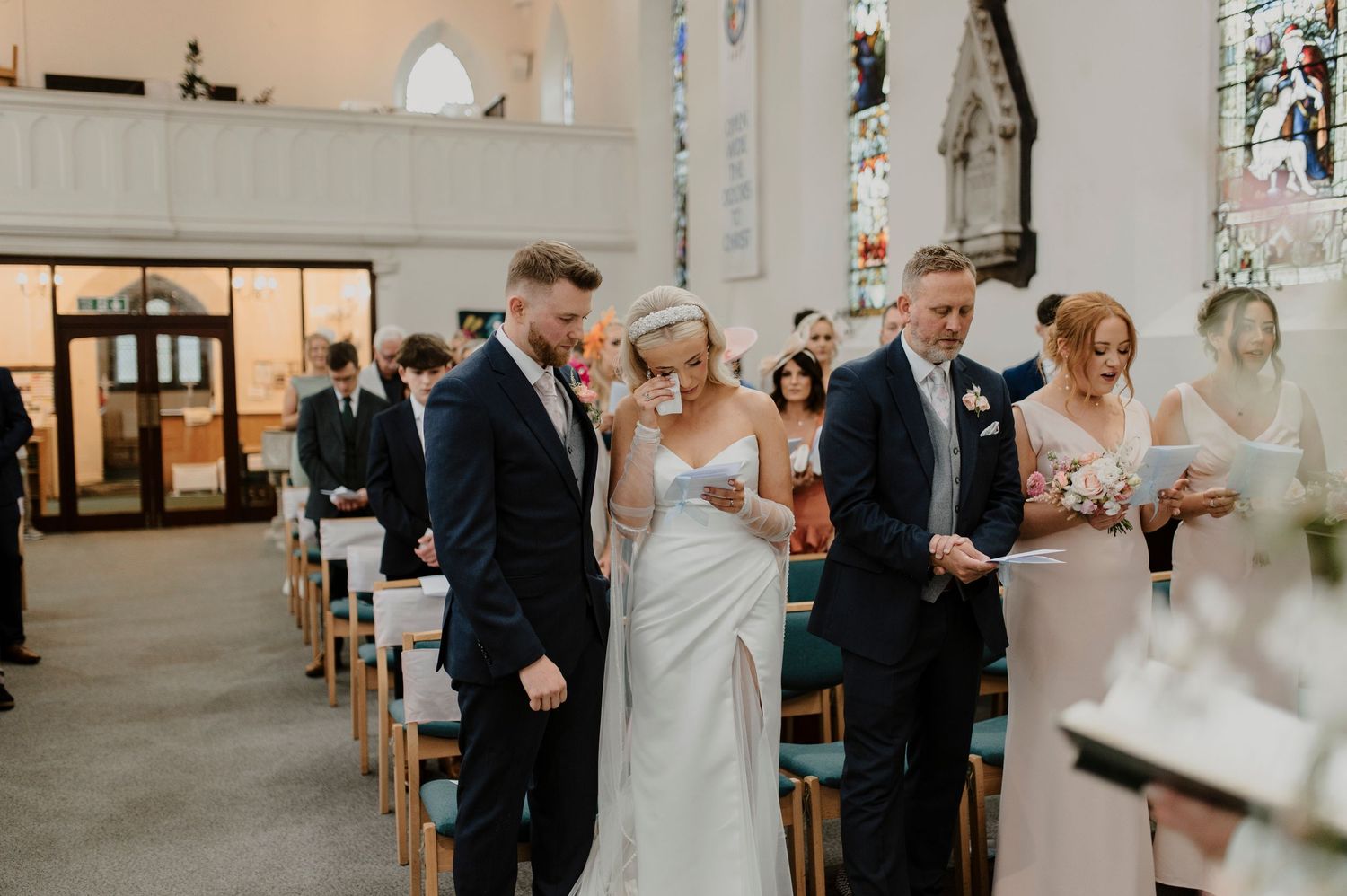 Wedding ceremony taking place inside a bright church with white walls and stained glass windows.