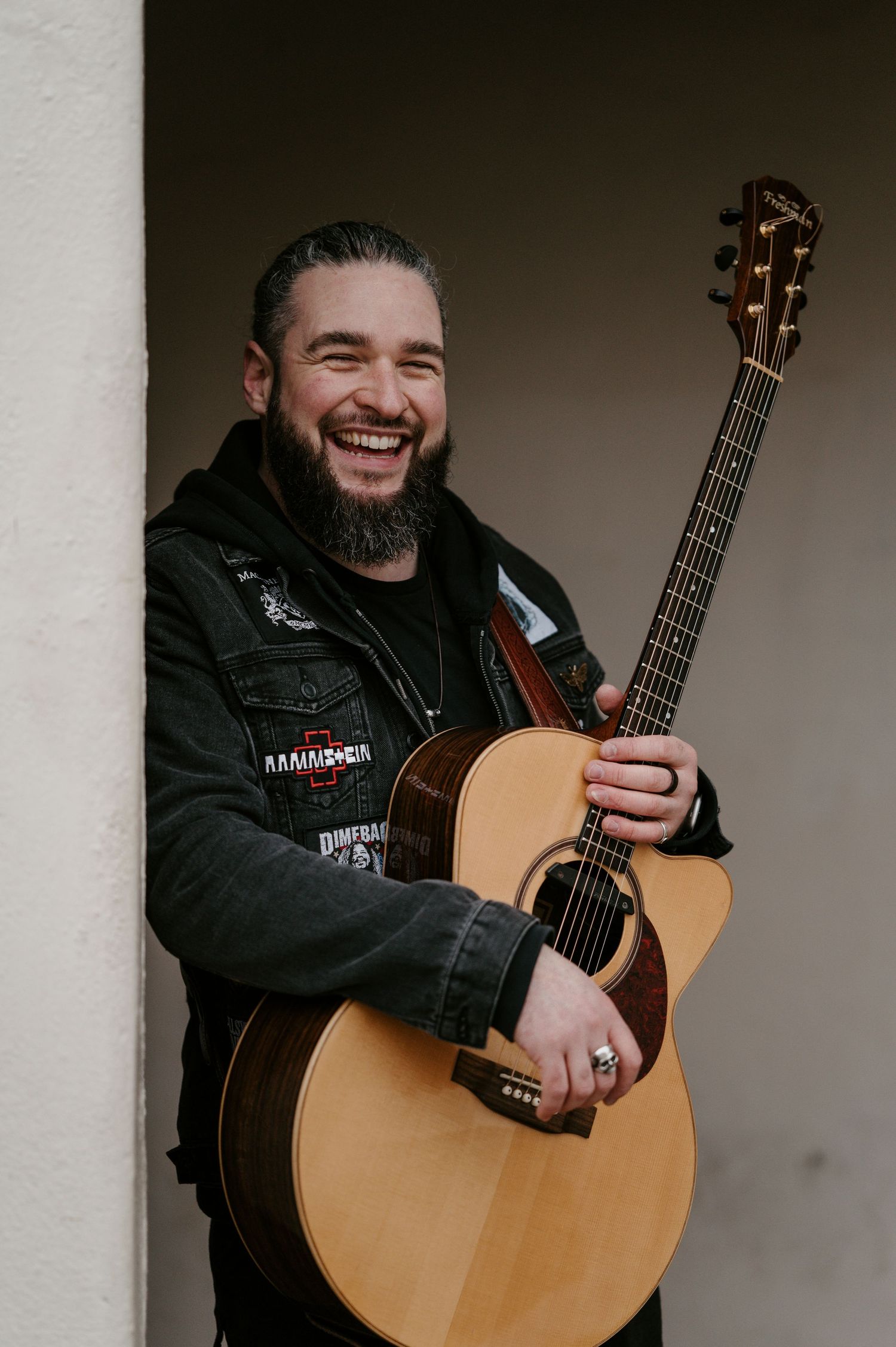 Musician in dark clothing plays acoustic guitar while seated against light colored background.