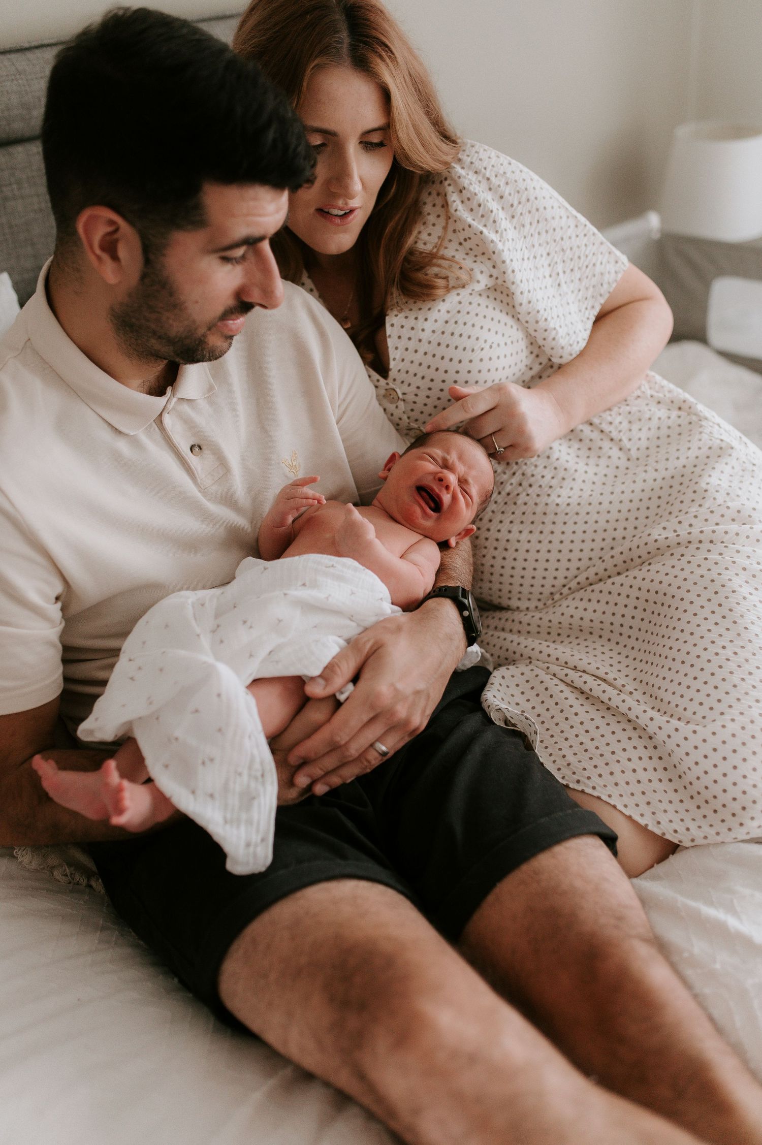A crying newborn baby wrapped in white cloth is held during an intimate family moment on a couch.