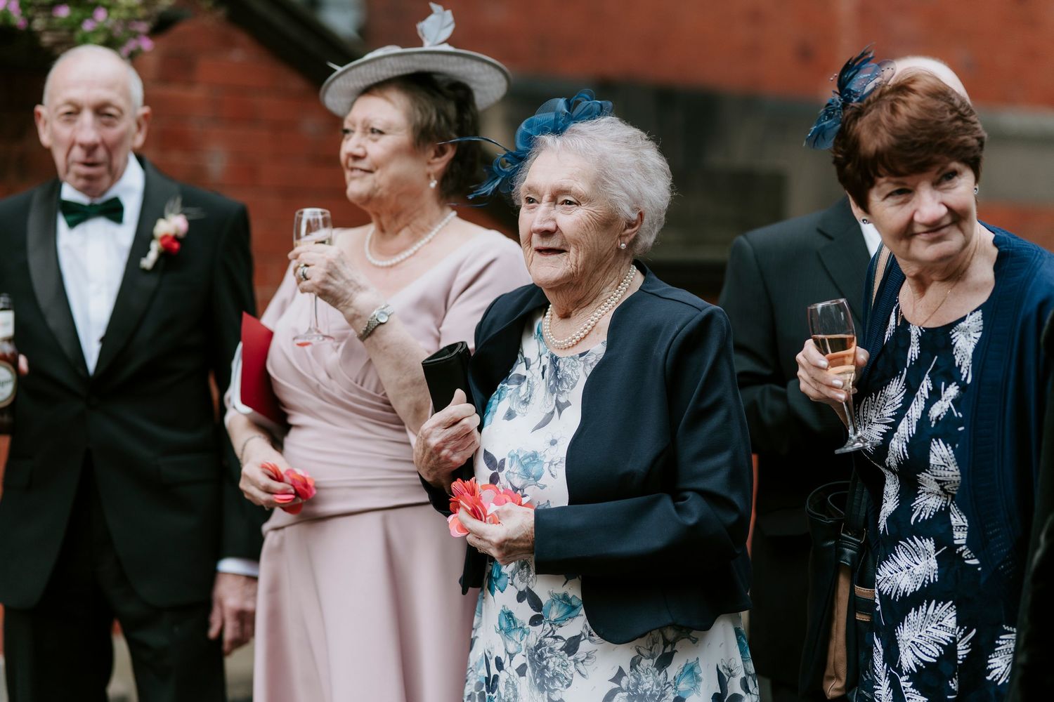 A group of wedding guests in formal attire laugh and smile together at an outdoor celebration.