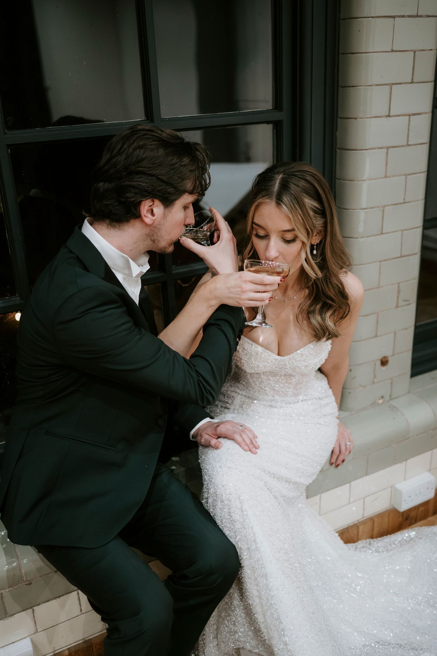 A couple shares a romantic moment on their wedding day as they sit together near a white tiled wall.