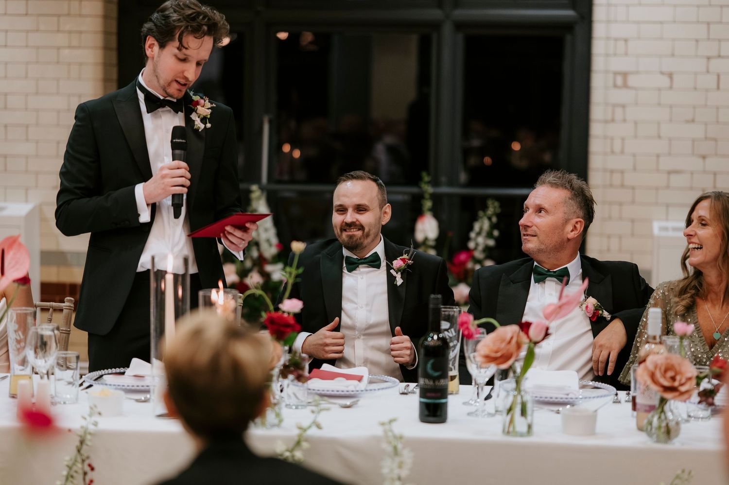 Wedding guests laugh and smile during dinner speeches at an elegant indoor reception.