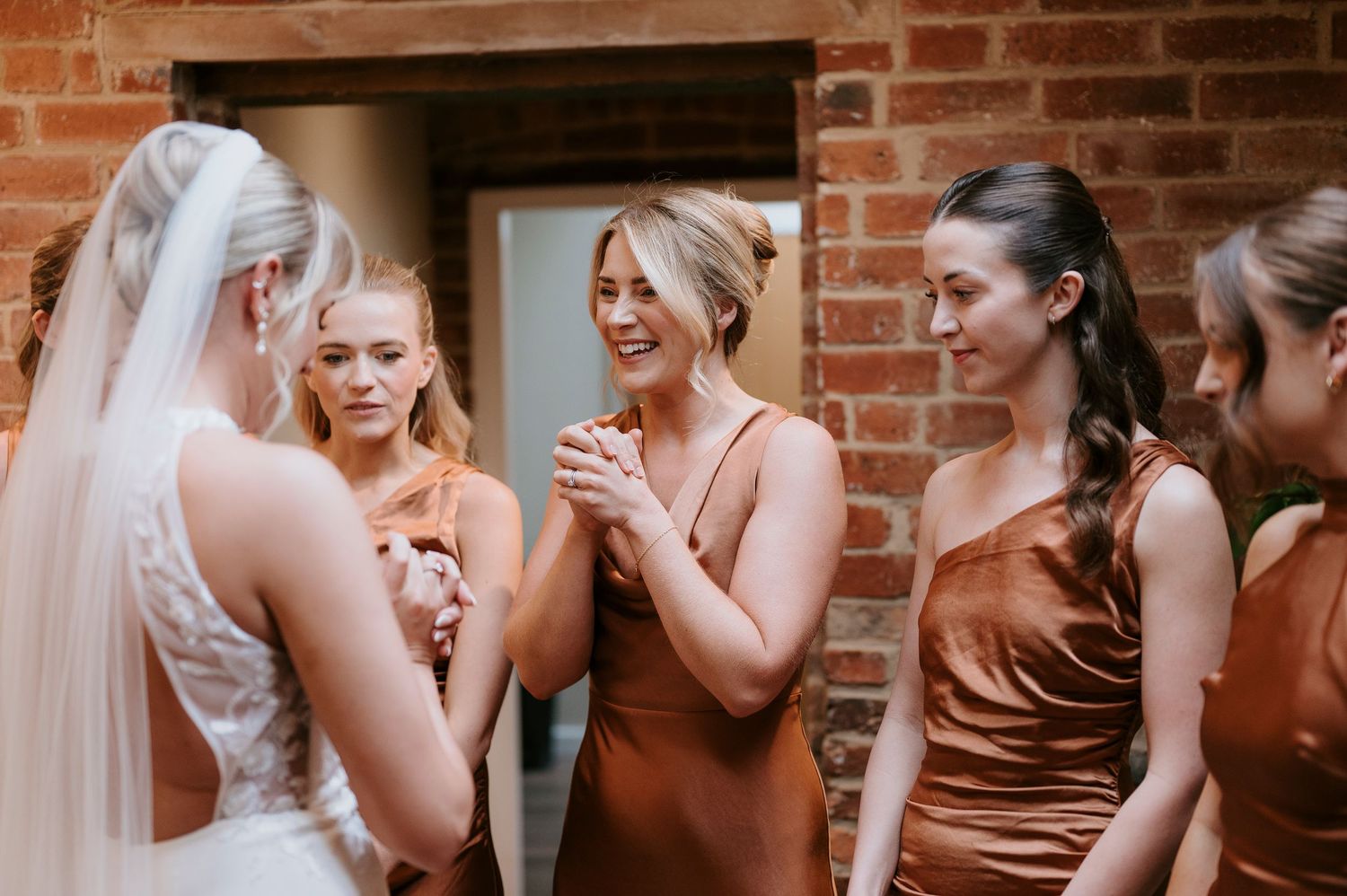 Bride in white wedding dress shares emotional moment with bridesmaids wearing rust-colored satin dresses against brick wall.